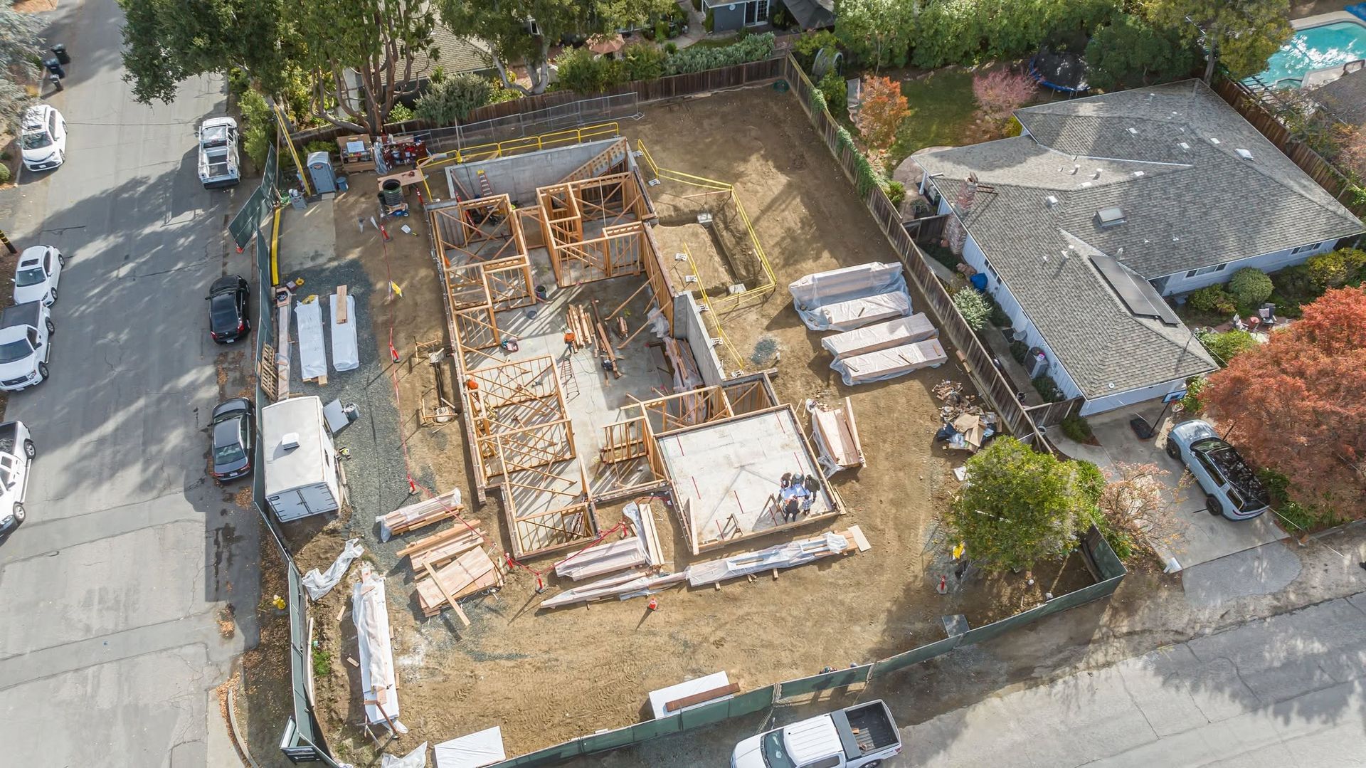 Aerial view of a construction site beside a road, with a partially built structure and nearby trees and houses