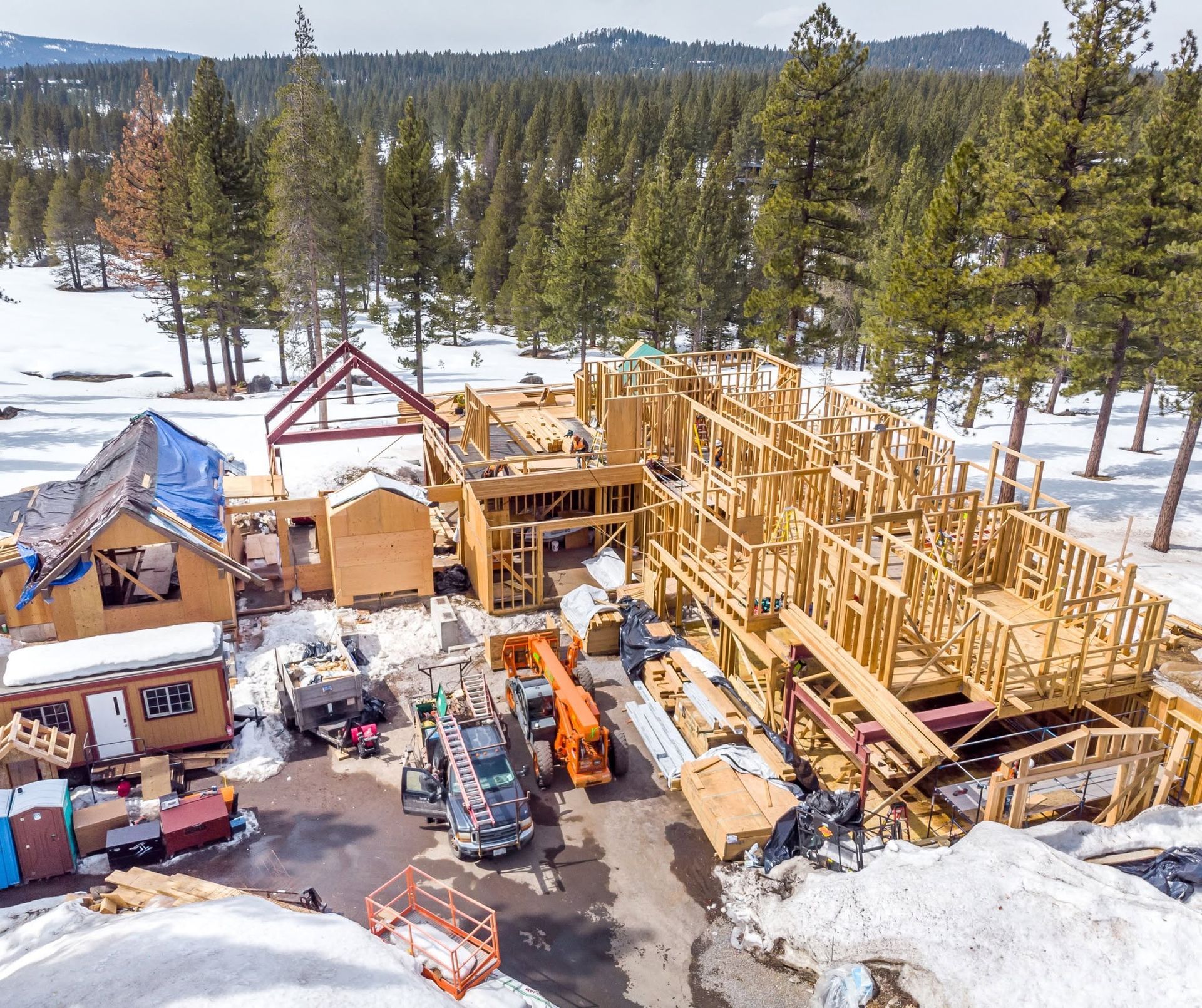Snowy construction site with wood framing, equipment, and pine trees in the background.