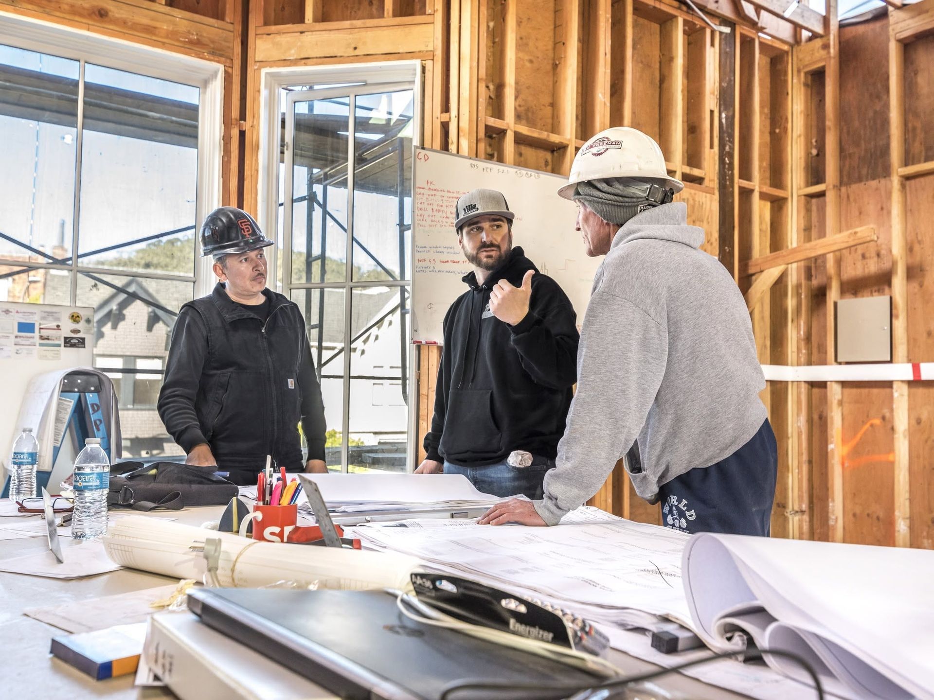 Three workers in hard hats review blueprints inside a framed construction site.