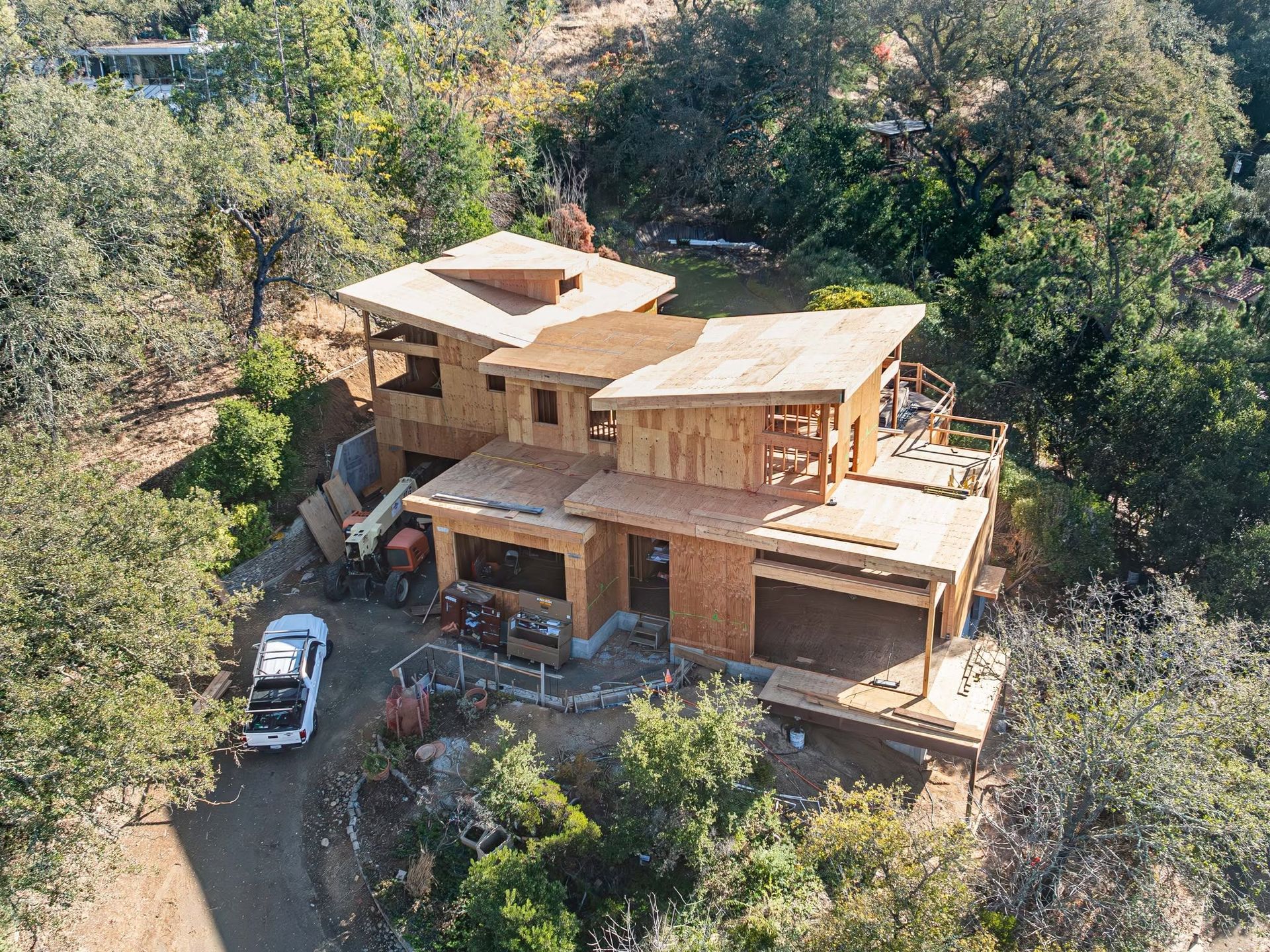 A modern wooden house under construction on a wooded hillside, with a car parked nearby.