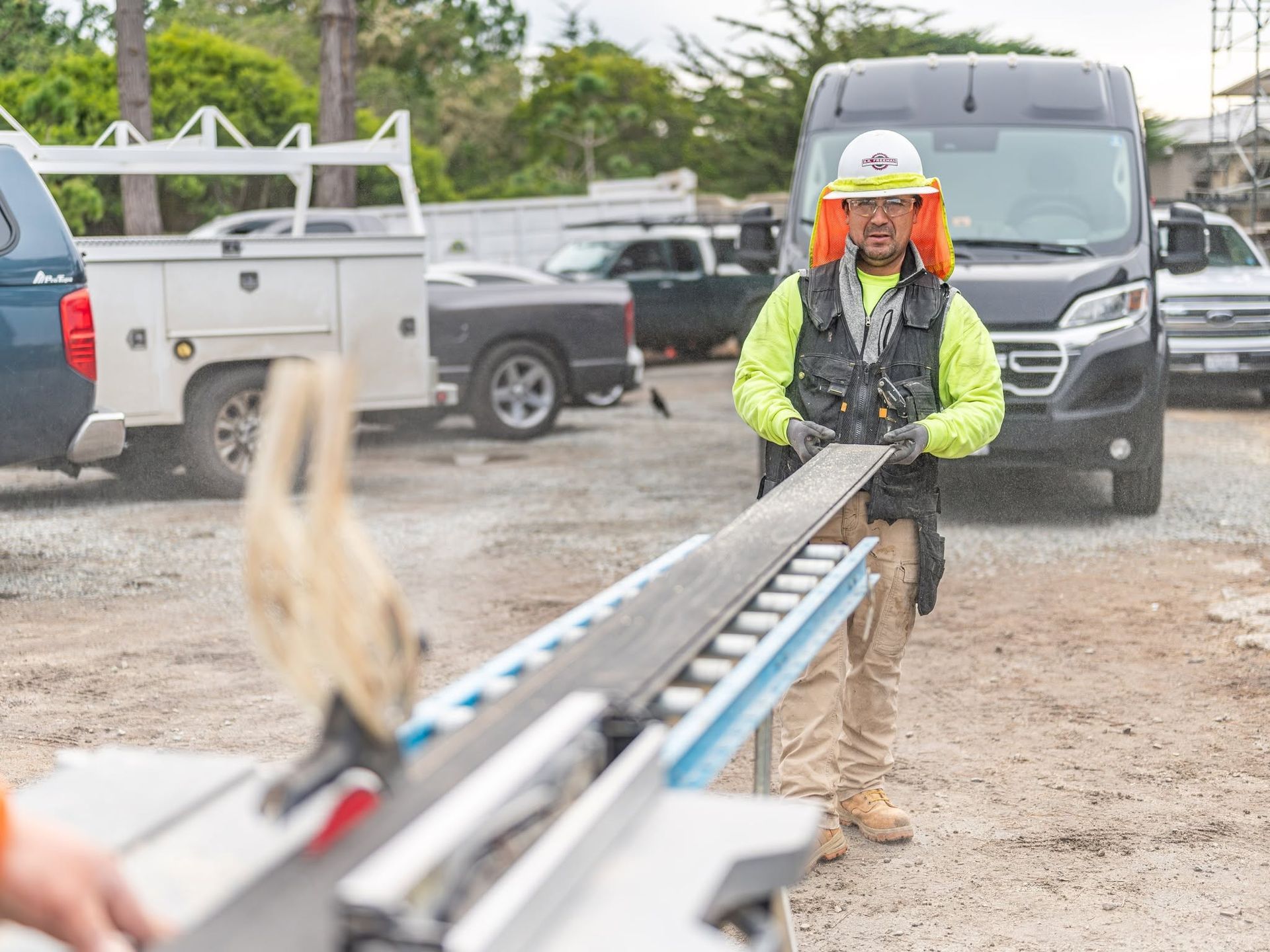 Construction worker in hi-vis gear guides a long beam near vans on a muddy worksite.
