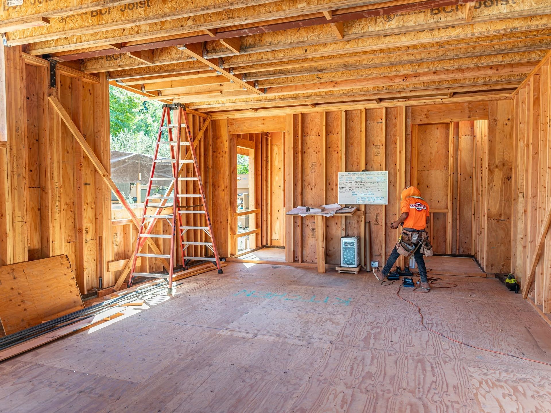 Wood-framed house interior under construction, with ladders, exposed beams, and a worker in an orange shirt.