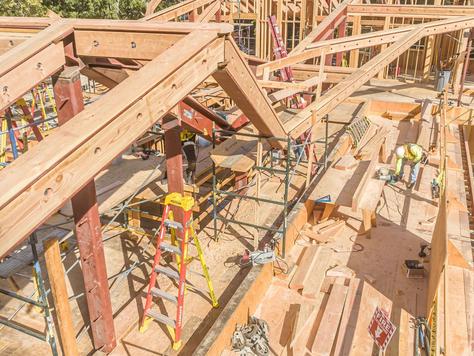 Construction workers framing a wooden roof with ladders and scattered lumber on a building site.