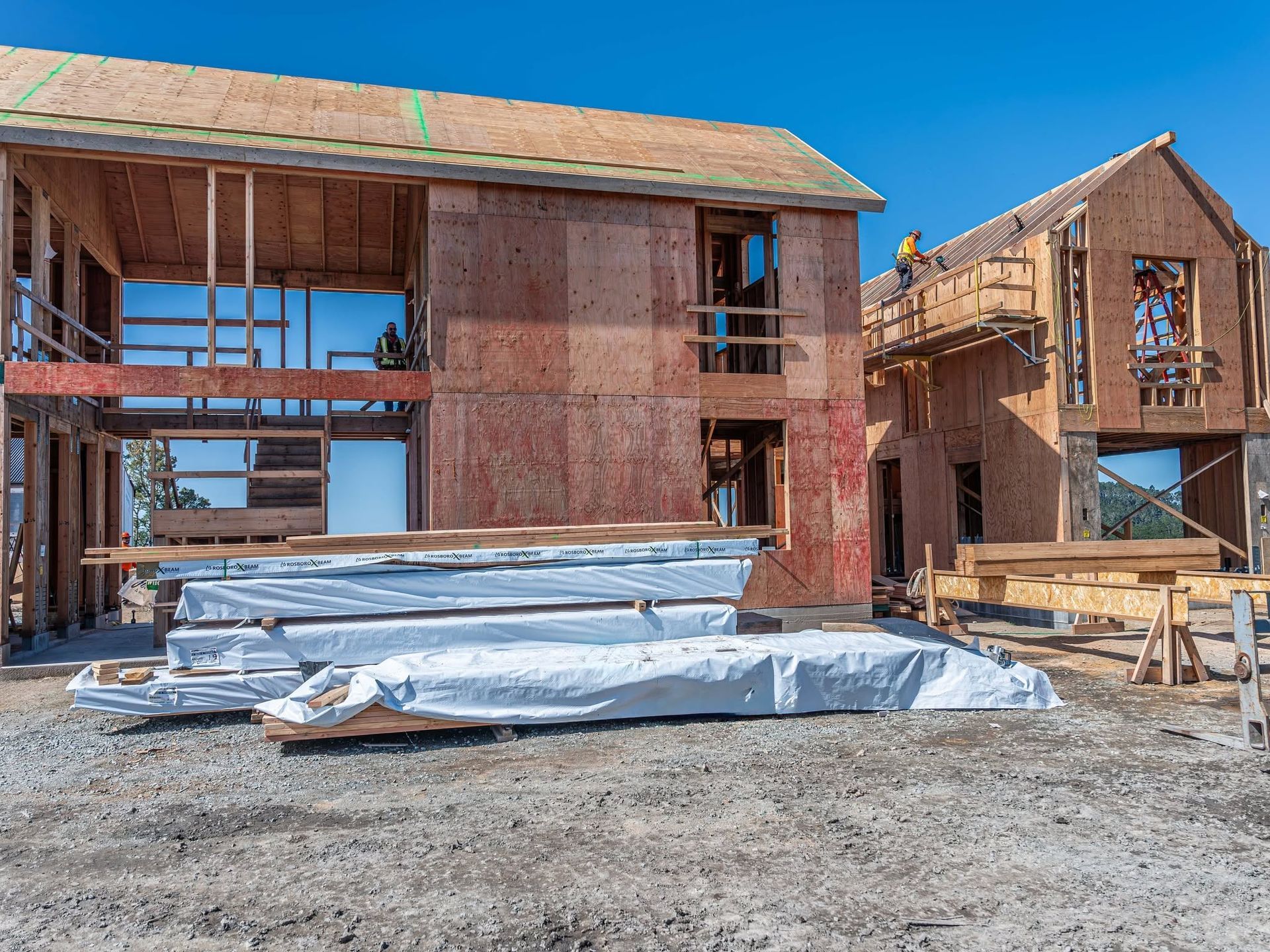 Two unfinished wooden houses under construction with wrapped materials on a dirt lot.