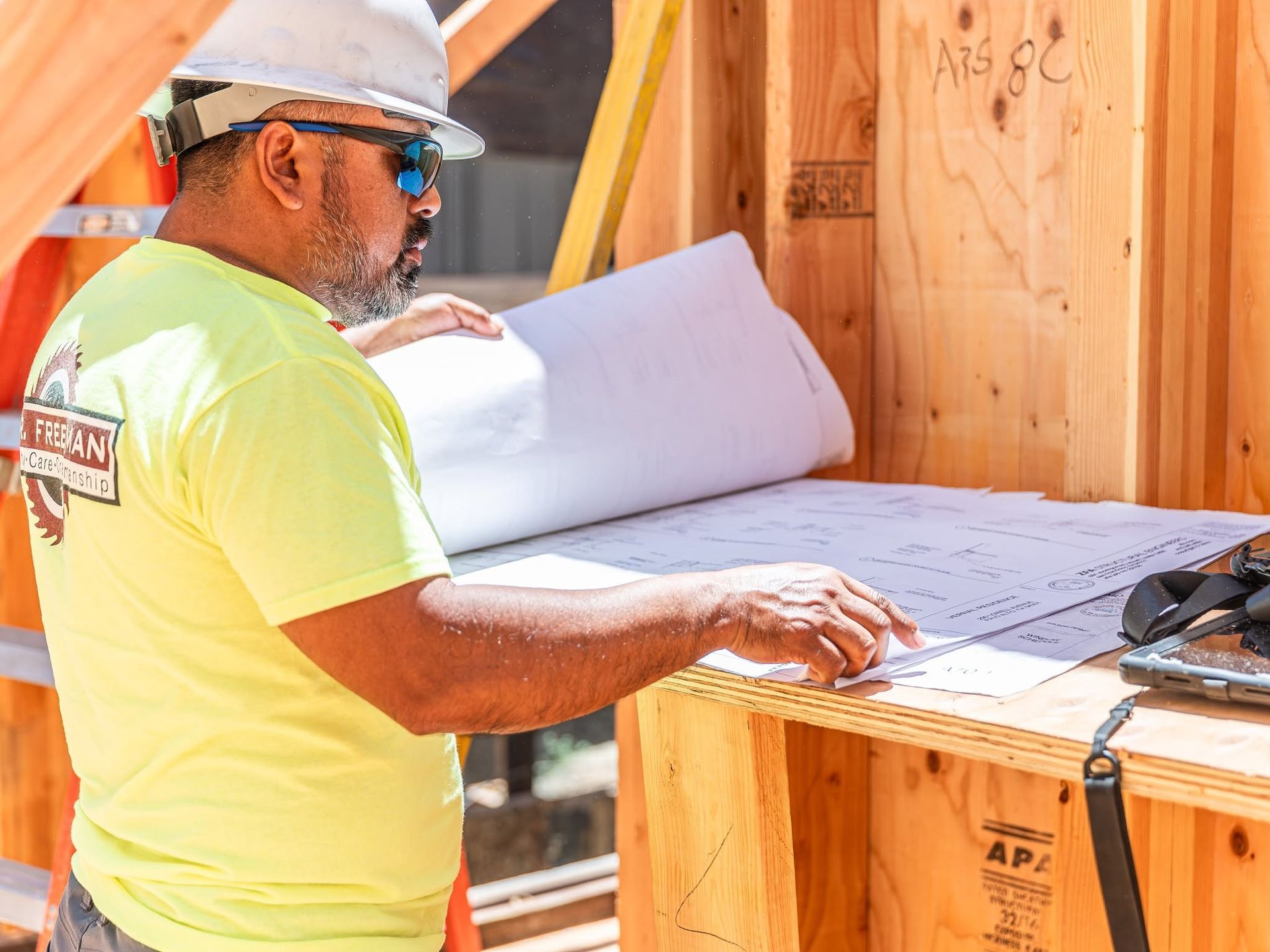 Construction worker in a hard hat reviews blueprints at a wooden framing site.