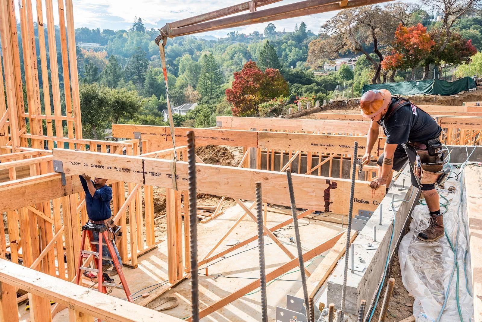 Construction workers building a wooden house frame on a hillside, with one worker on a ladder.