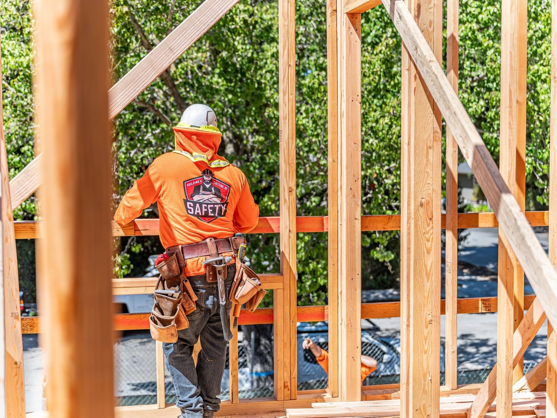 Construction worker in orange jacket and hard hat framing a wooden building structure outdoors