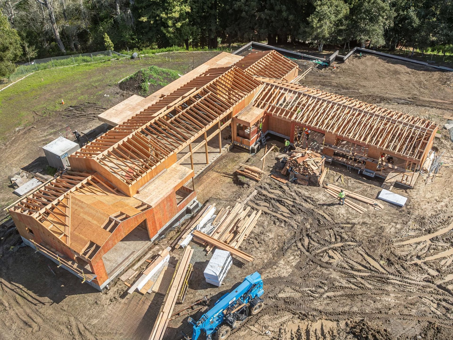 Wooden house frame under construction on a dirt lot with blue machinery nearby