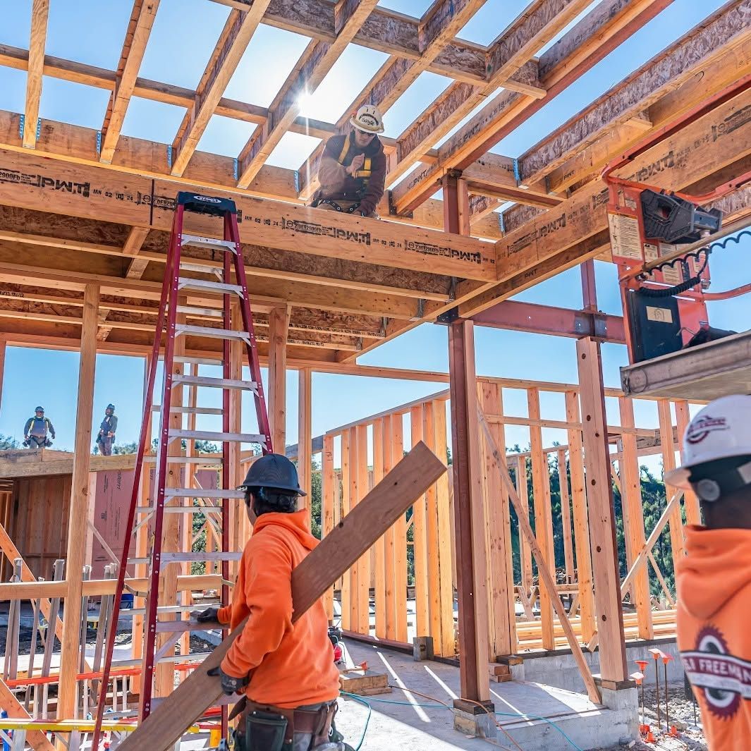 Construction workers framing a wooden building, with ladders and exposed roof beams on a sunny site