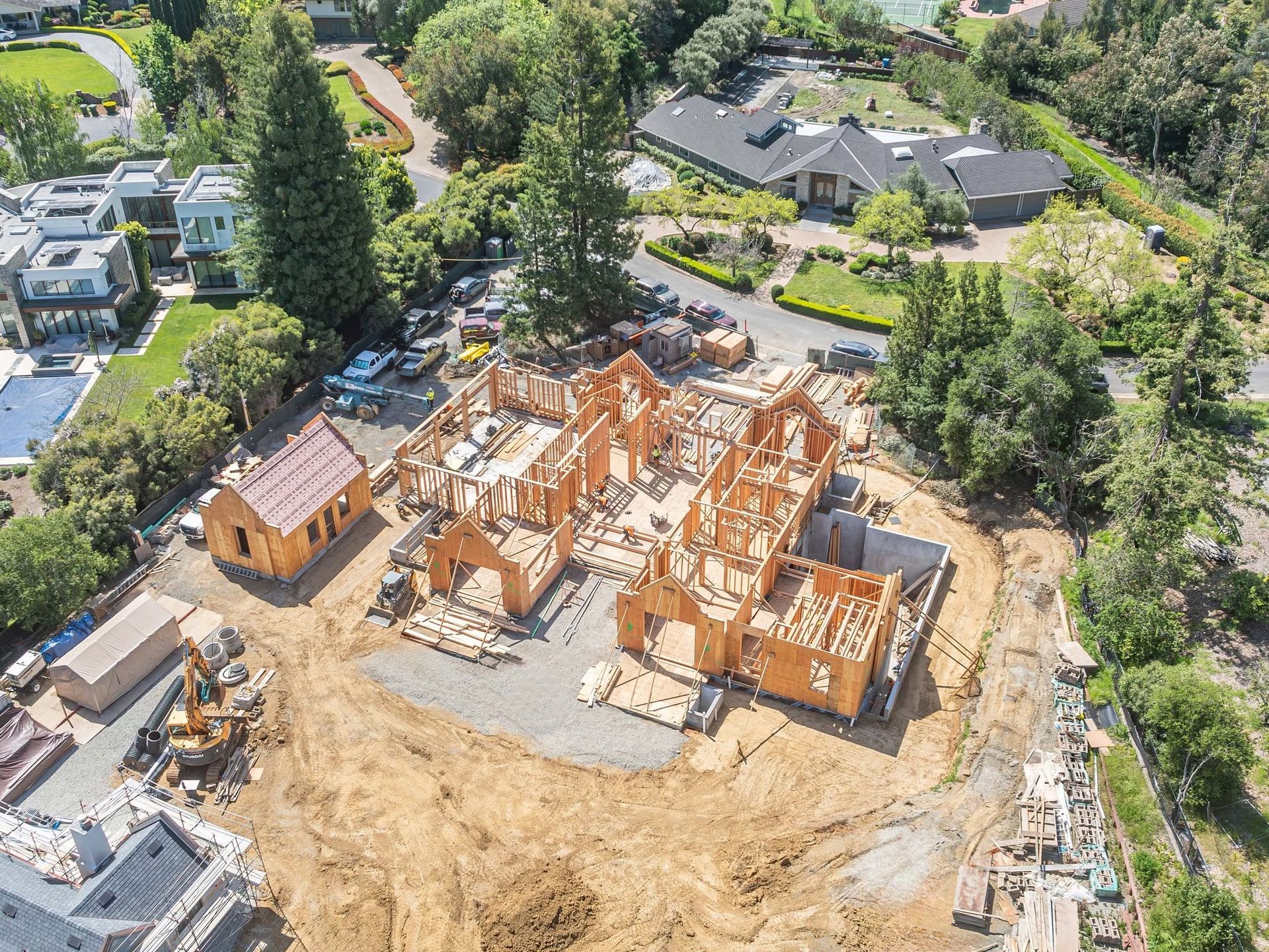 Aerial view of a house under construction with wood framing and nearby completed homes and trees.
