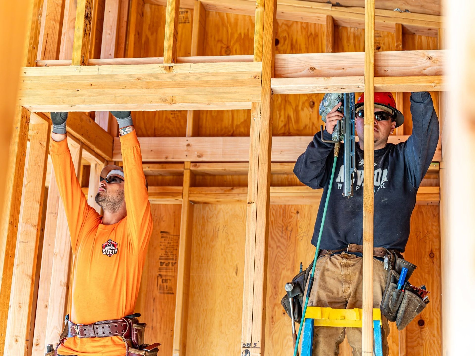 Two construction workers in orange and navy shirts install wooden framing inside a house