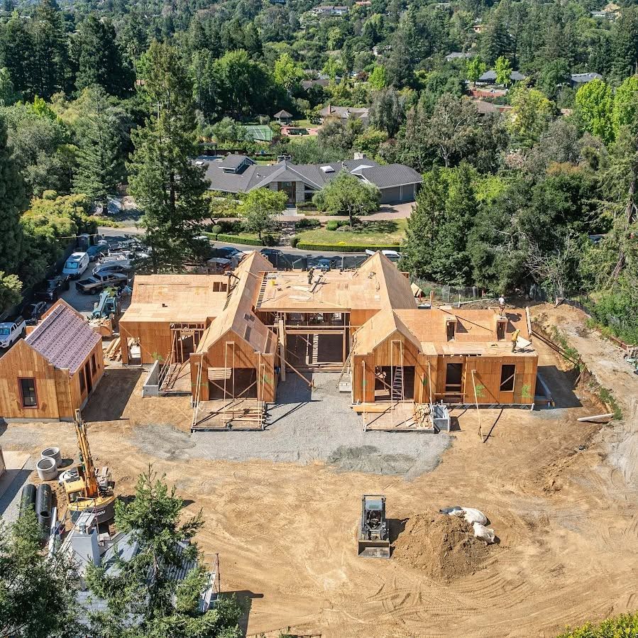 Aerial view of a wooden house under construction on a cleared lot, surrounded by trees.