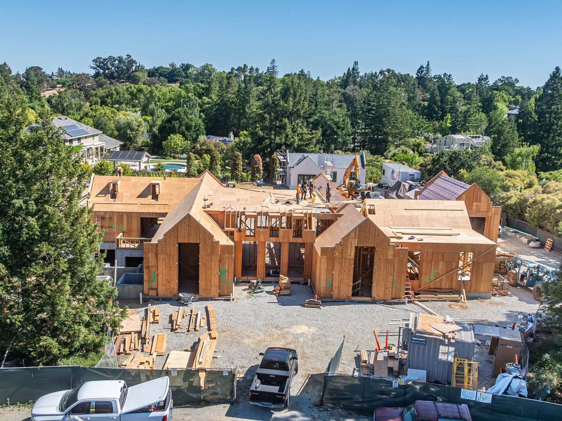 Wood-framed houses under construction on a residential lot with trees and parked vehicles nearby