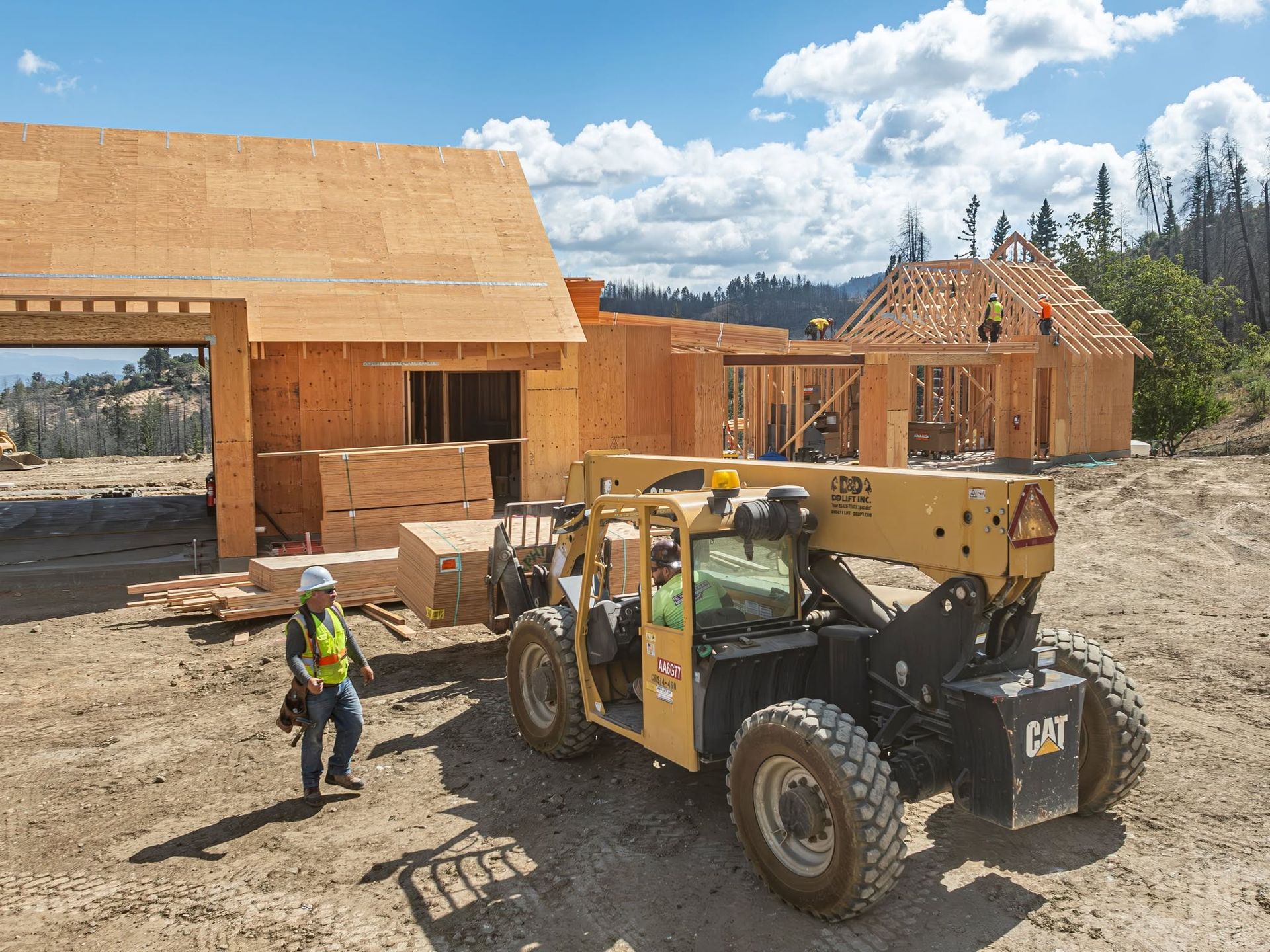 Construction site with workers and a yellow telehandler beside unfinished wooden buildings under a blue sky