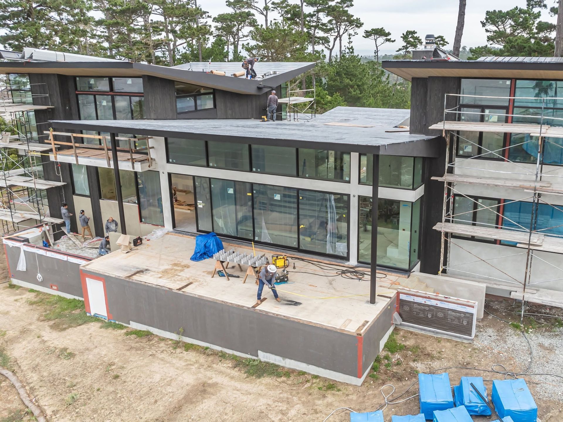 Modern house under construction with workers, large glass windows, and blue tarps on the site.