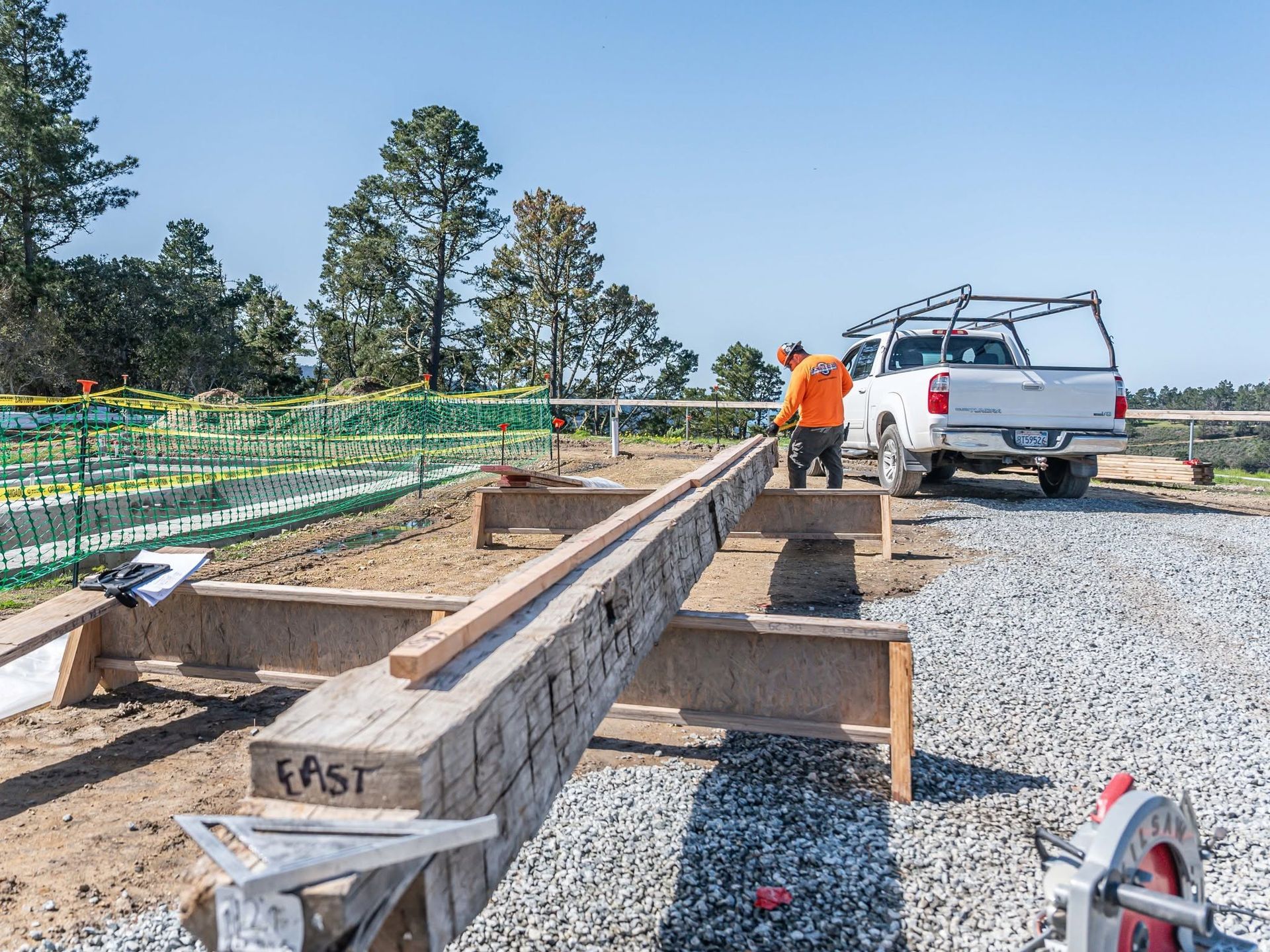 Construction workers beside long wooden beams at a gravel site, with a white truck and trees in the background