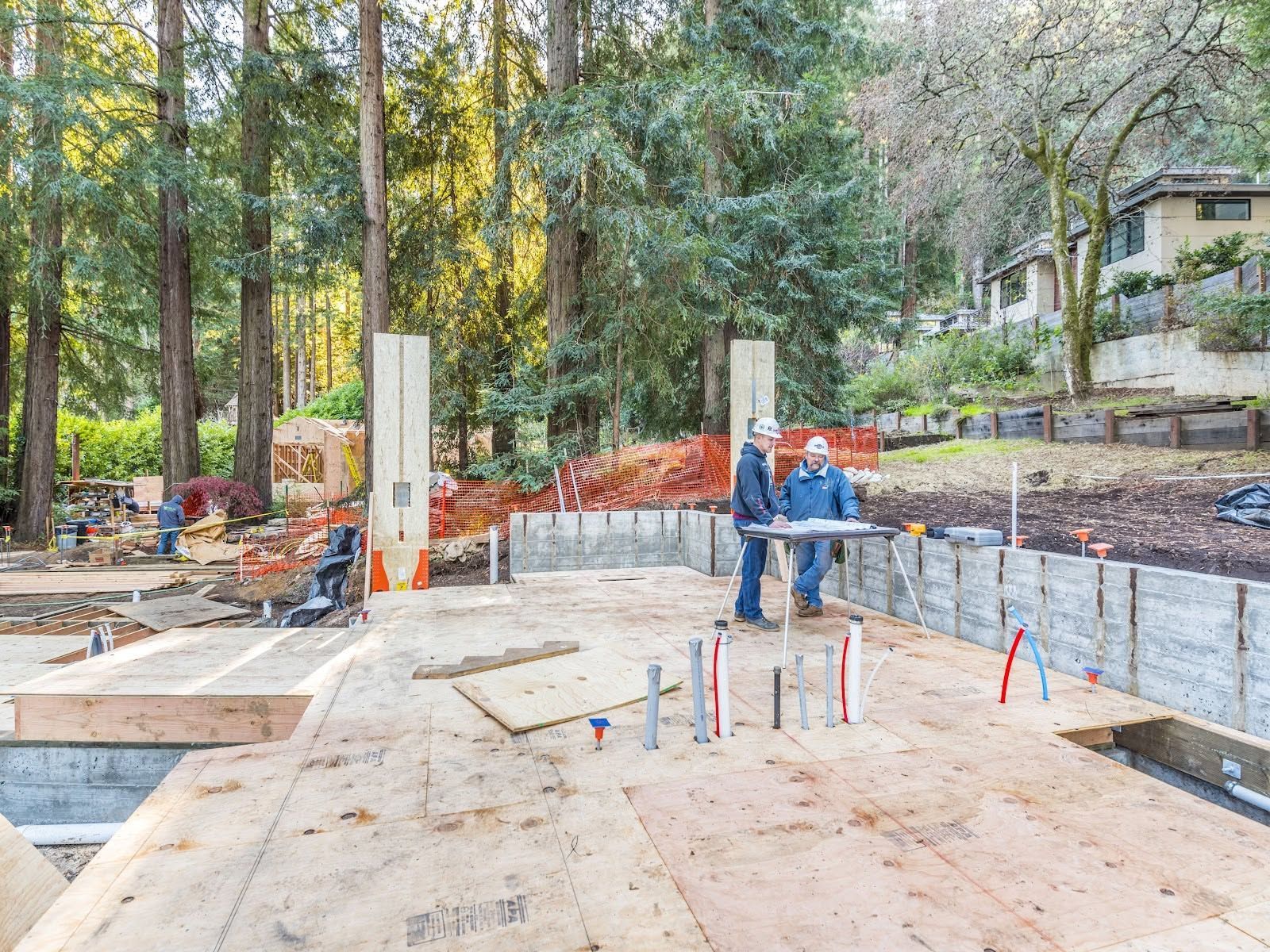 Construction site with concrete foundation, workers, and orange safety fencing in a wooded residential area