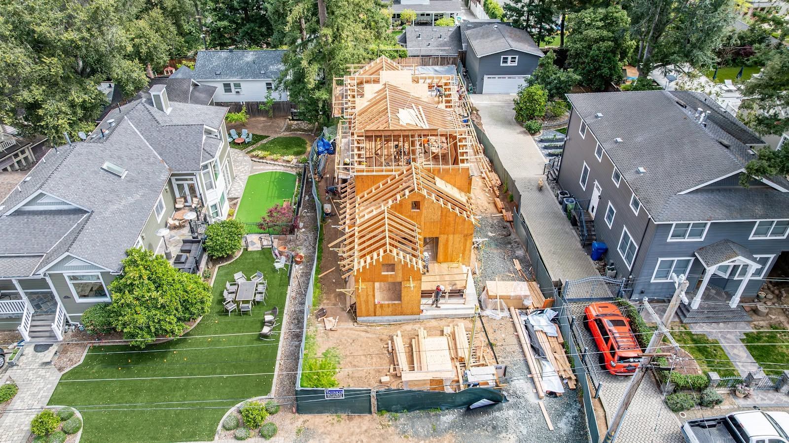 Aerial view of a house under construction between finished homes, with exposed wooden framing and parked vehicles nearby.