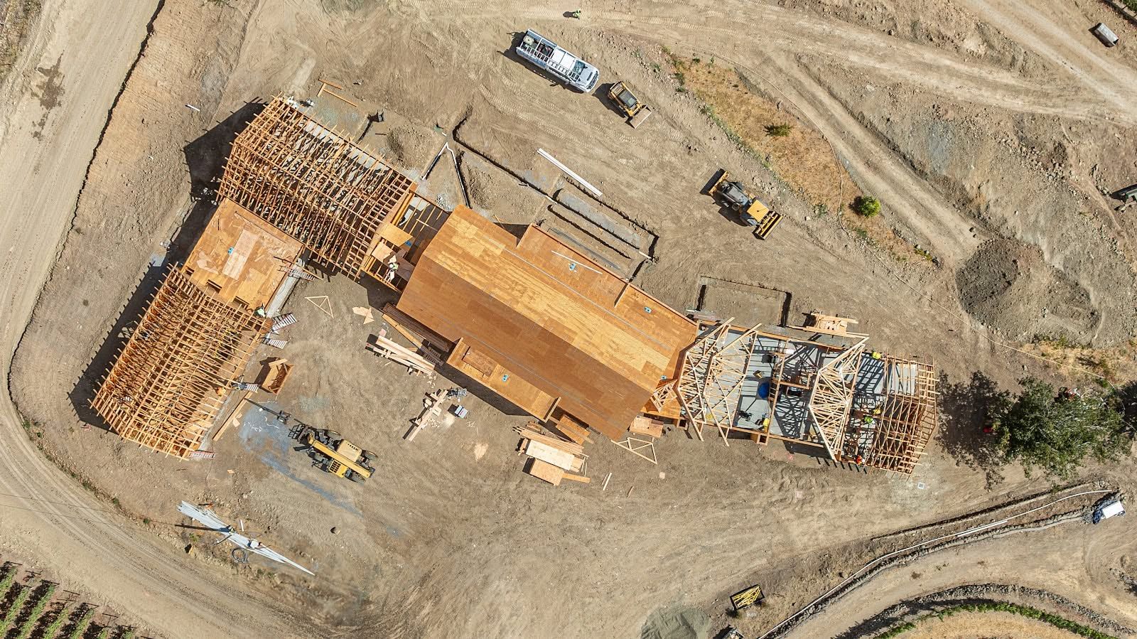Aerial view of a construction site with a long building, materials, and several vehicles on dirt ground