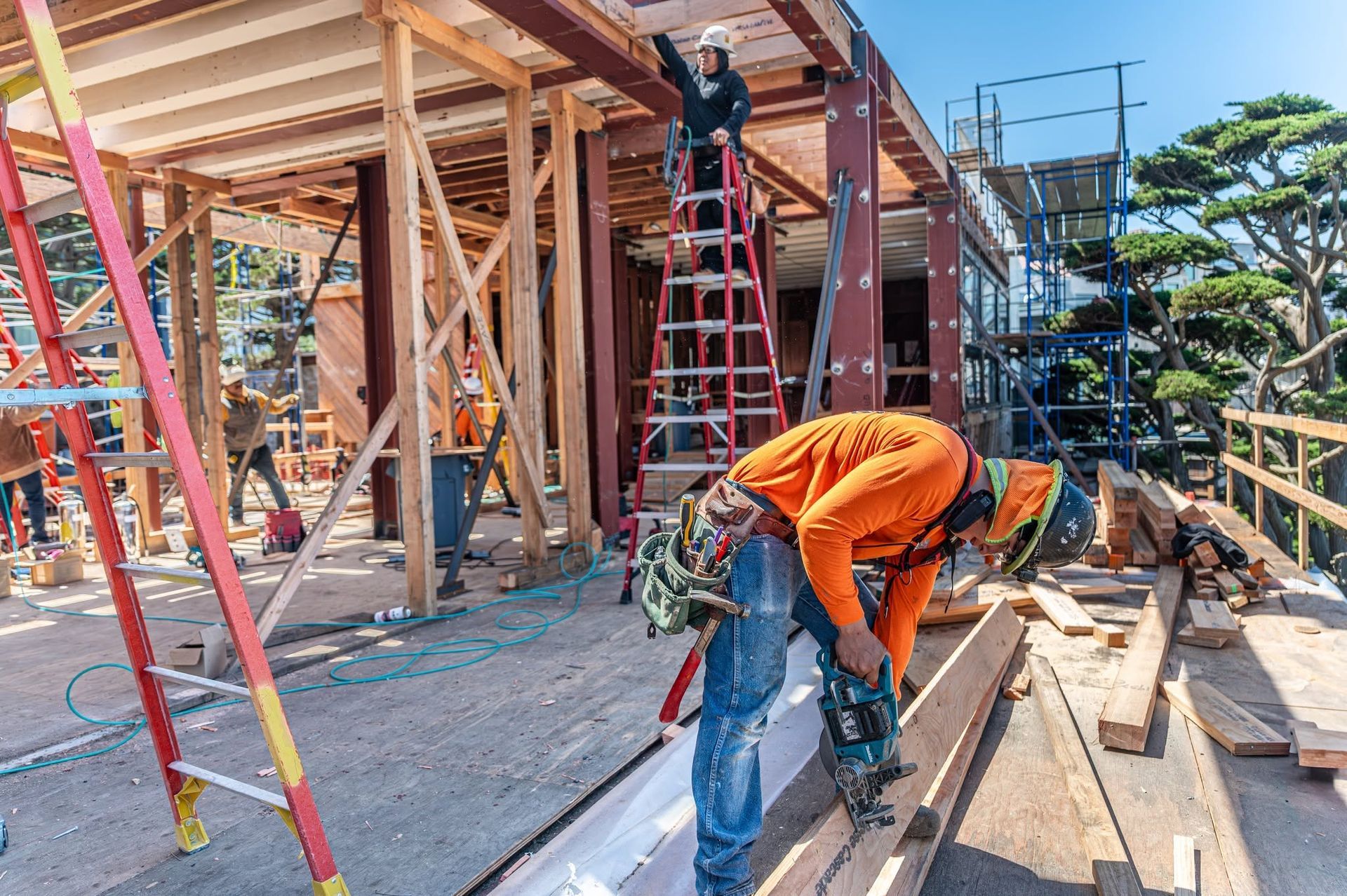 Construction workers on a building site, one in orange bending over tools near lumber and ladders.