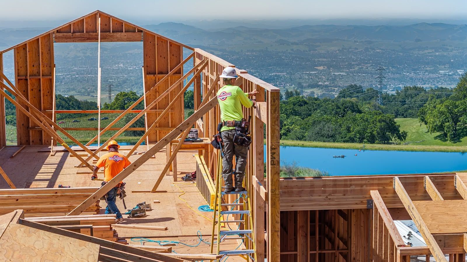 Construction workers framing a house with a scenic hillside and blue pool in the background
