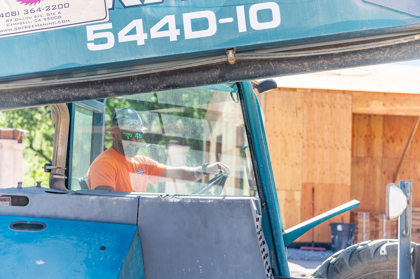 Worker inside a blue tractor cab with “544D-10” on the canopy, parked near a wooden wall