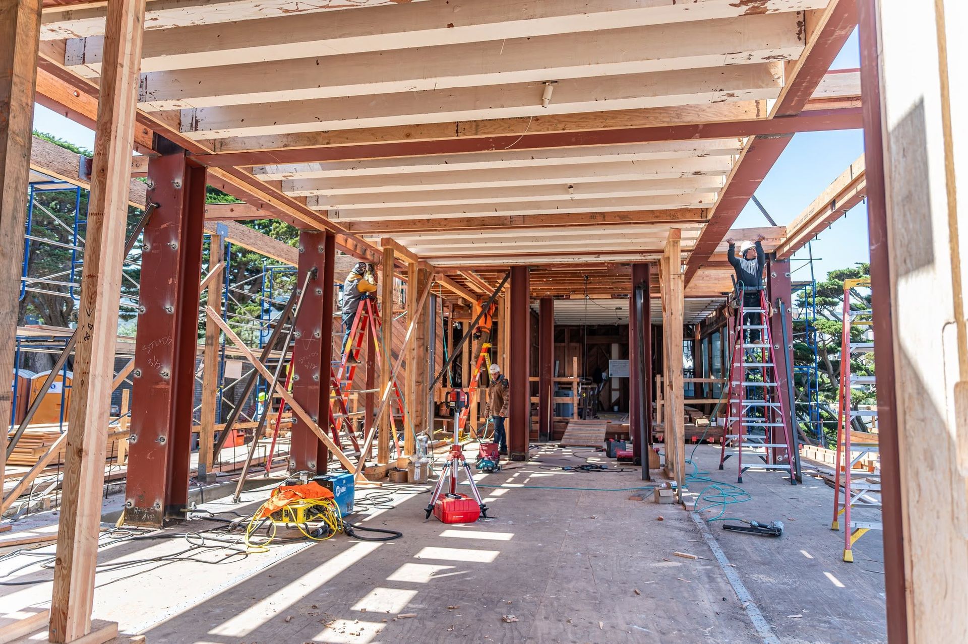 Wood-framed building under construction, with beams, scaffolding, and tools in a sunny open space