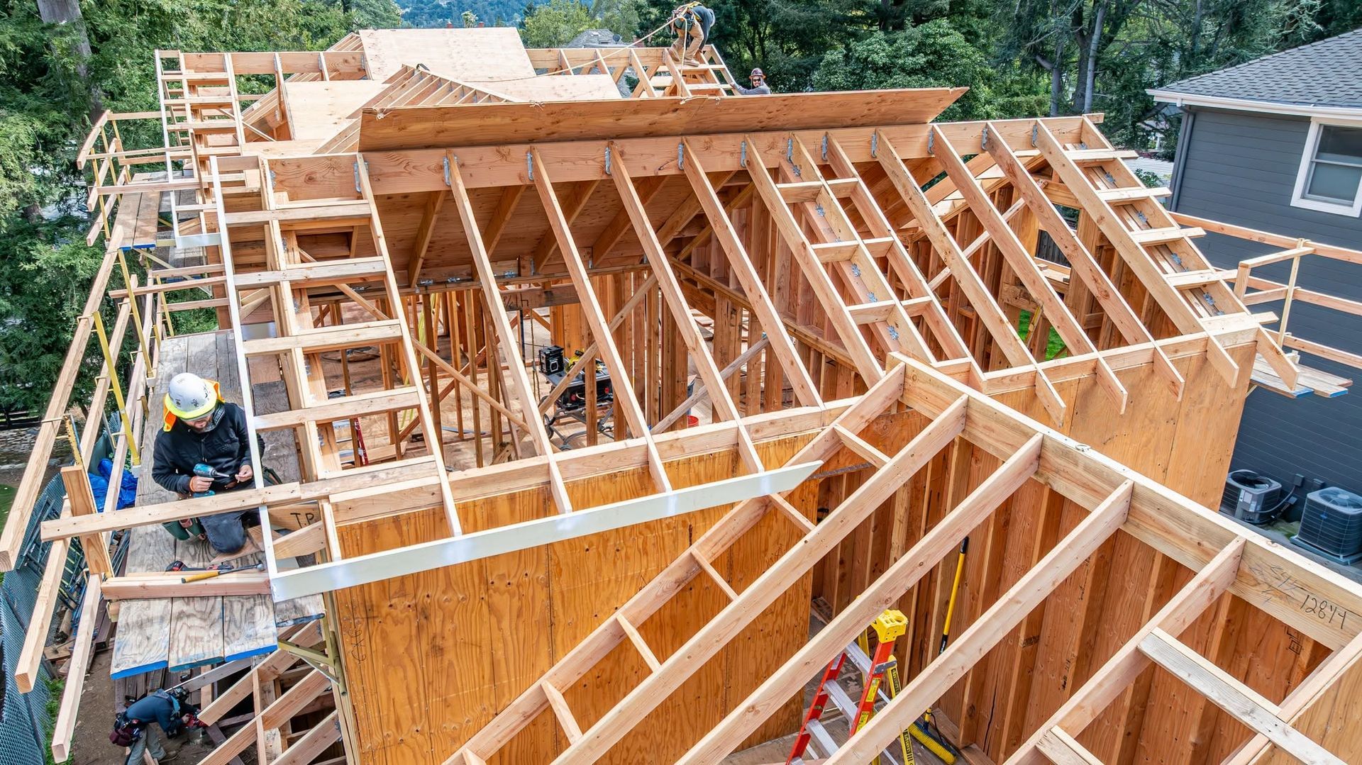 Wooden roof framing under construction with workers and ladders on a house site