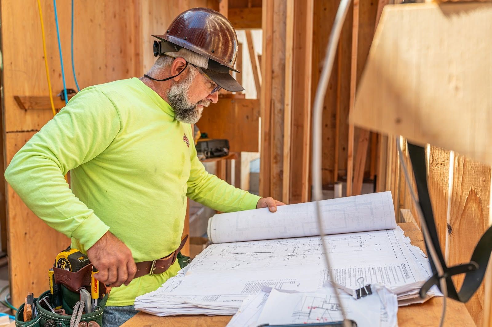 Construction worker in neon shirt reviewing blueprints inside a wooden-framed building