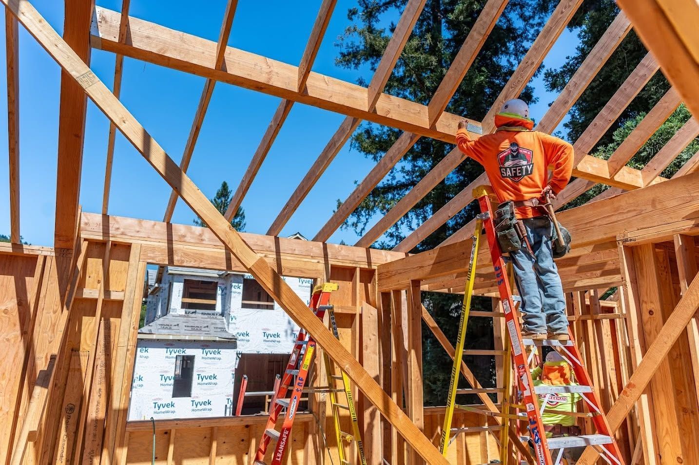 Construction workers framing a wooden house, one standing on a ladder under a bright blue sky.