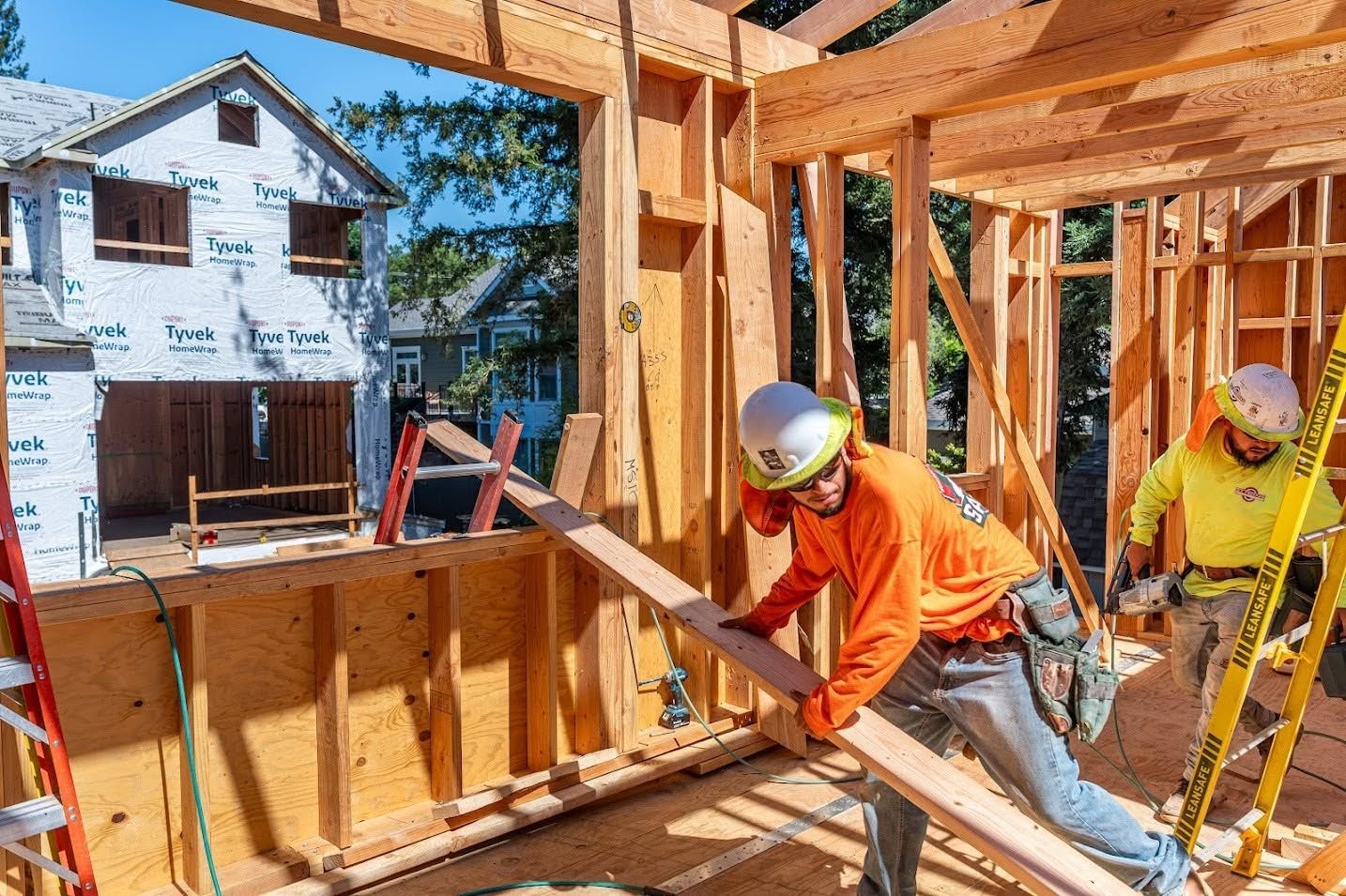 Construction workers framing a wooden house, one in orange and one in yellow, on a bright day