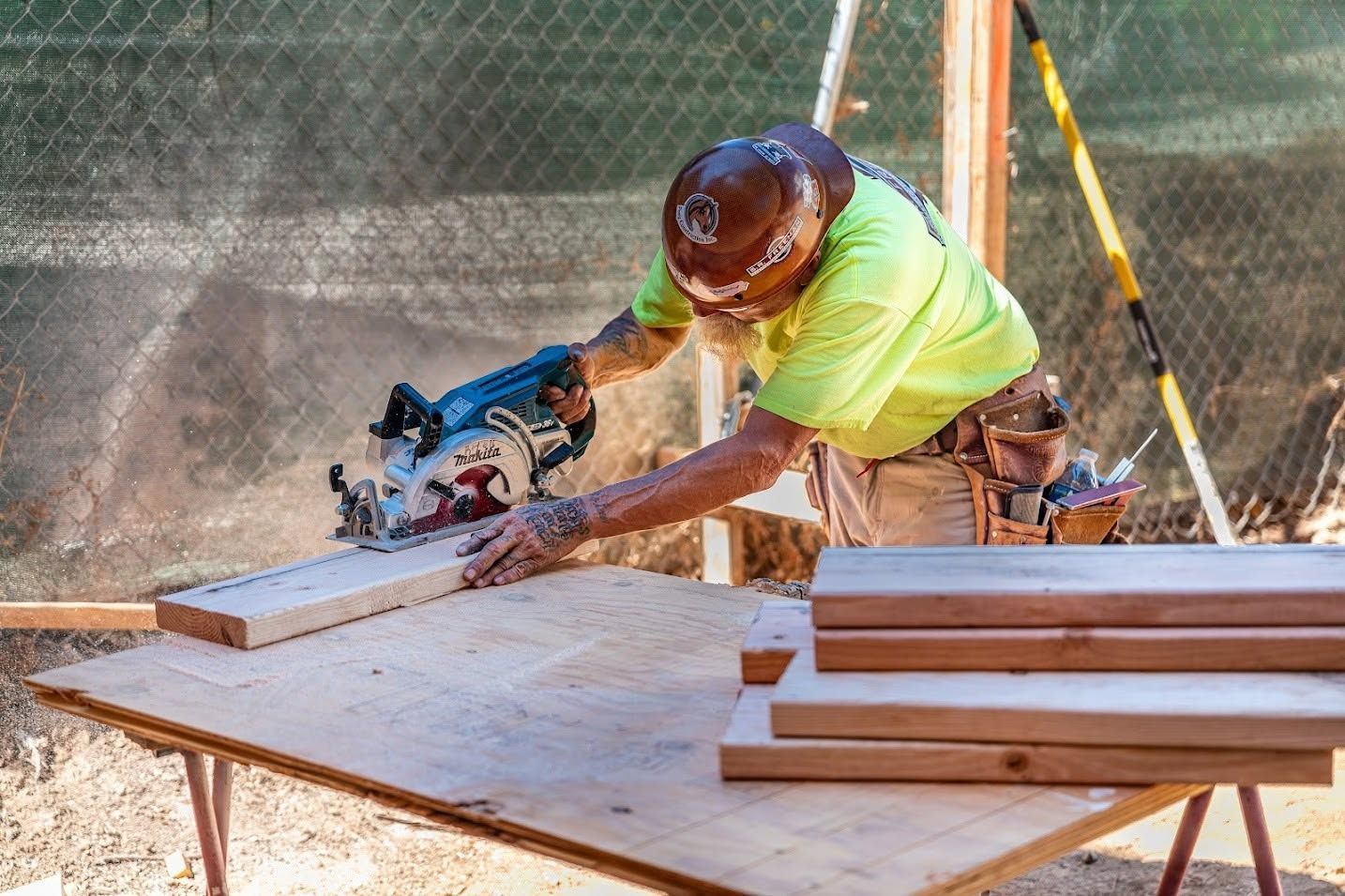 Worker cutting lumber with a circular saw at an outdoor worksite, with stacked wood nearby.