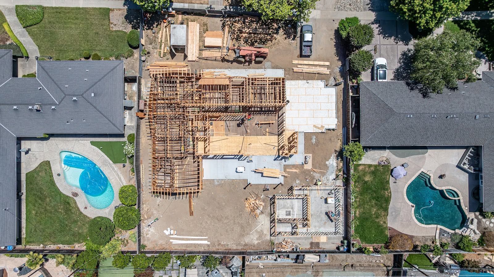Aerial view of a house under construction between two homes with backyard swimming pools.