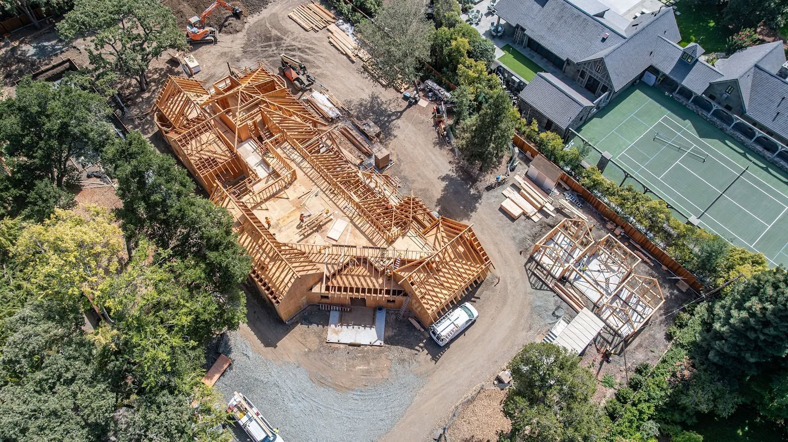 Aerial view of a house under construction beside another building and tennis courts, with exposed wooden framing.