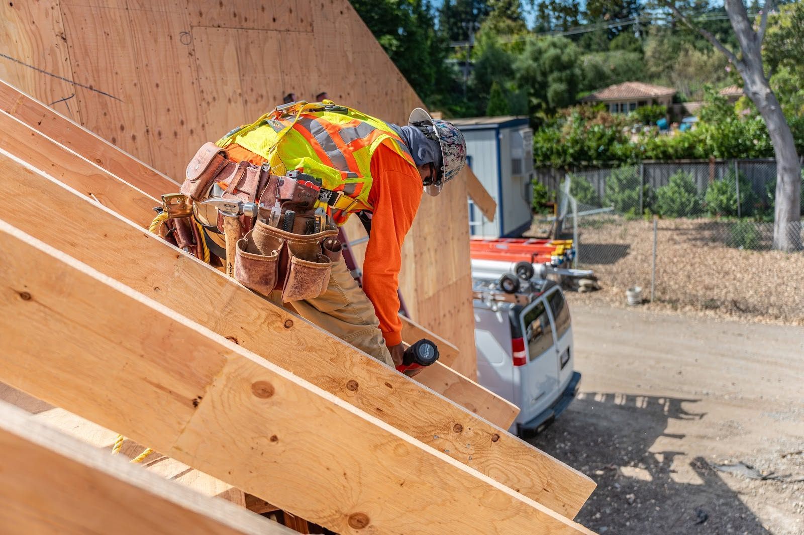 Construction worker in orange safety gear on a sloped wooden roof beside a work truck.