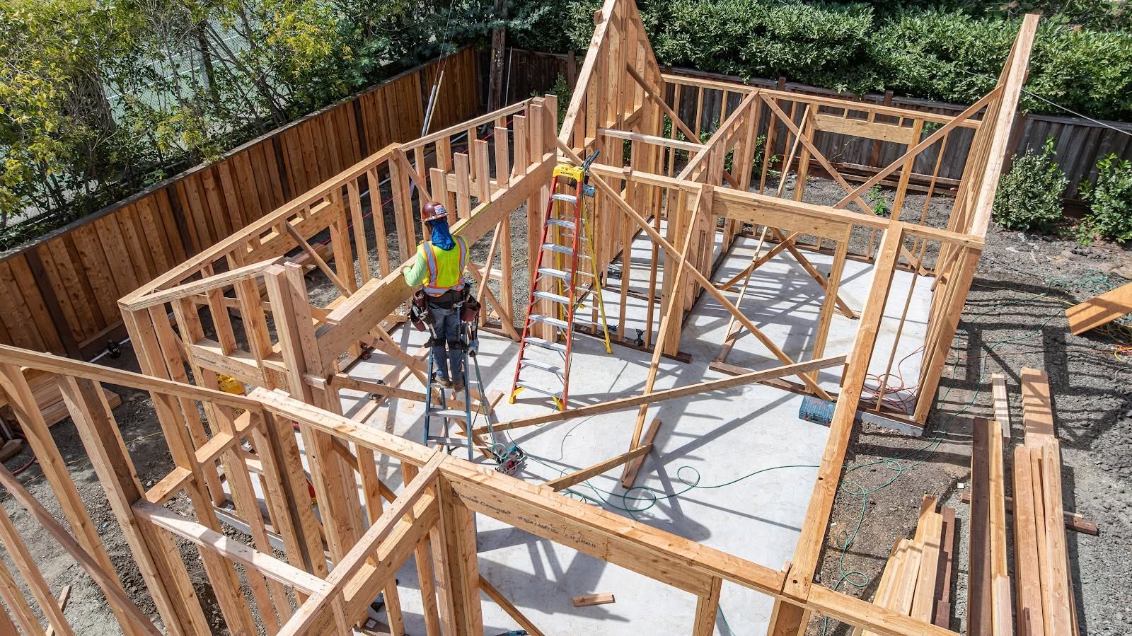 Wooden house framing under construction with workers and a white floor slab in sunlight