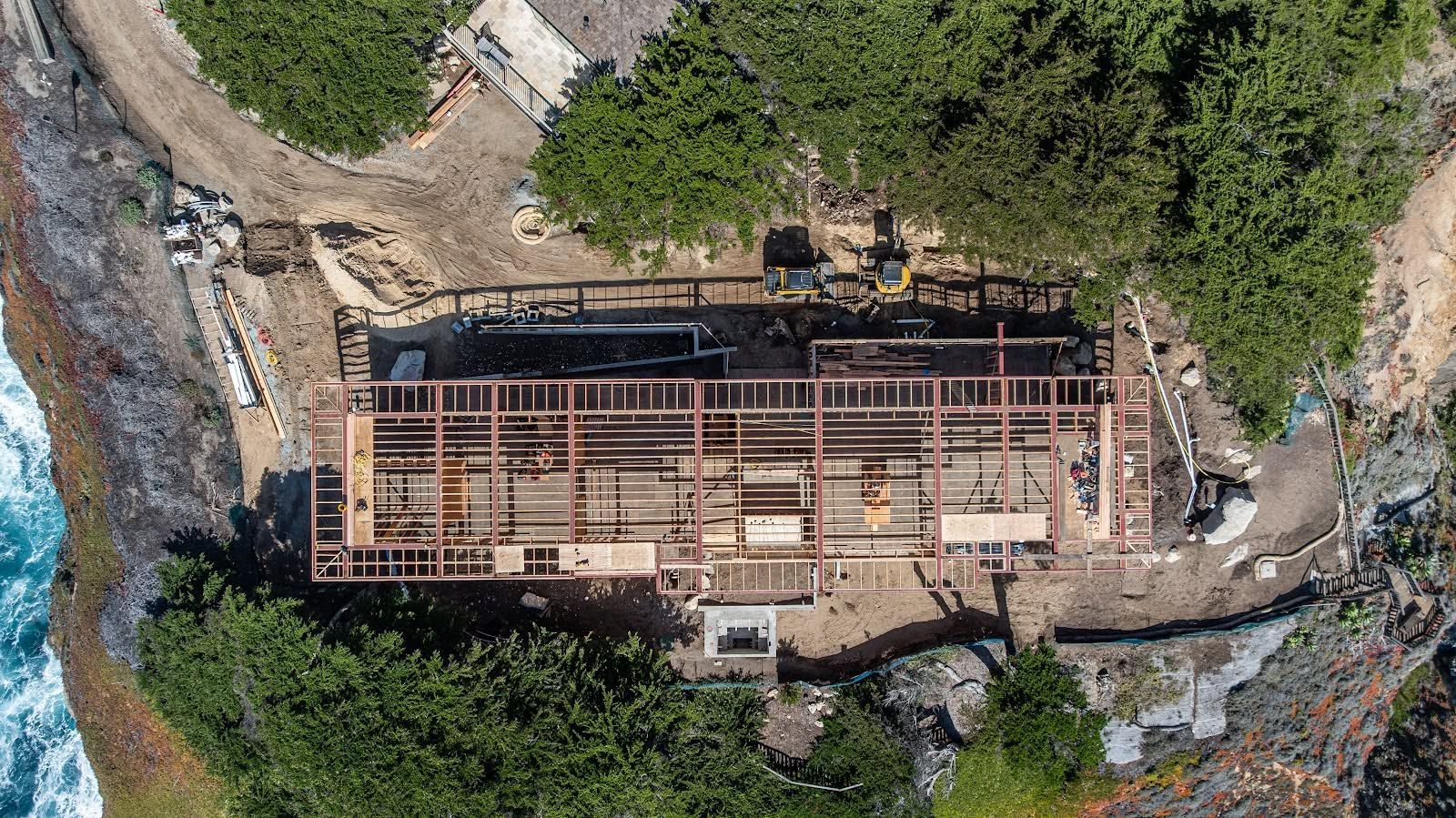 Aerial view of a rectangular construction site with exposed framing surrounded by dense green trees.