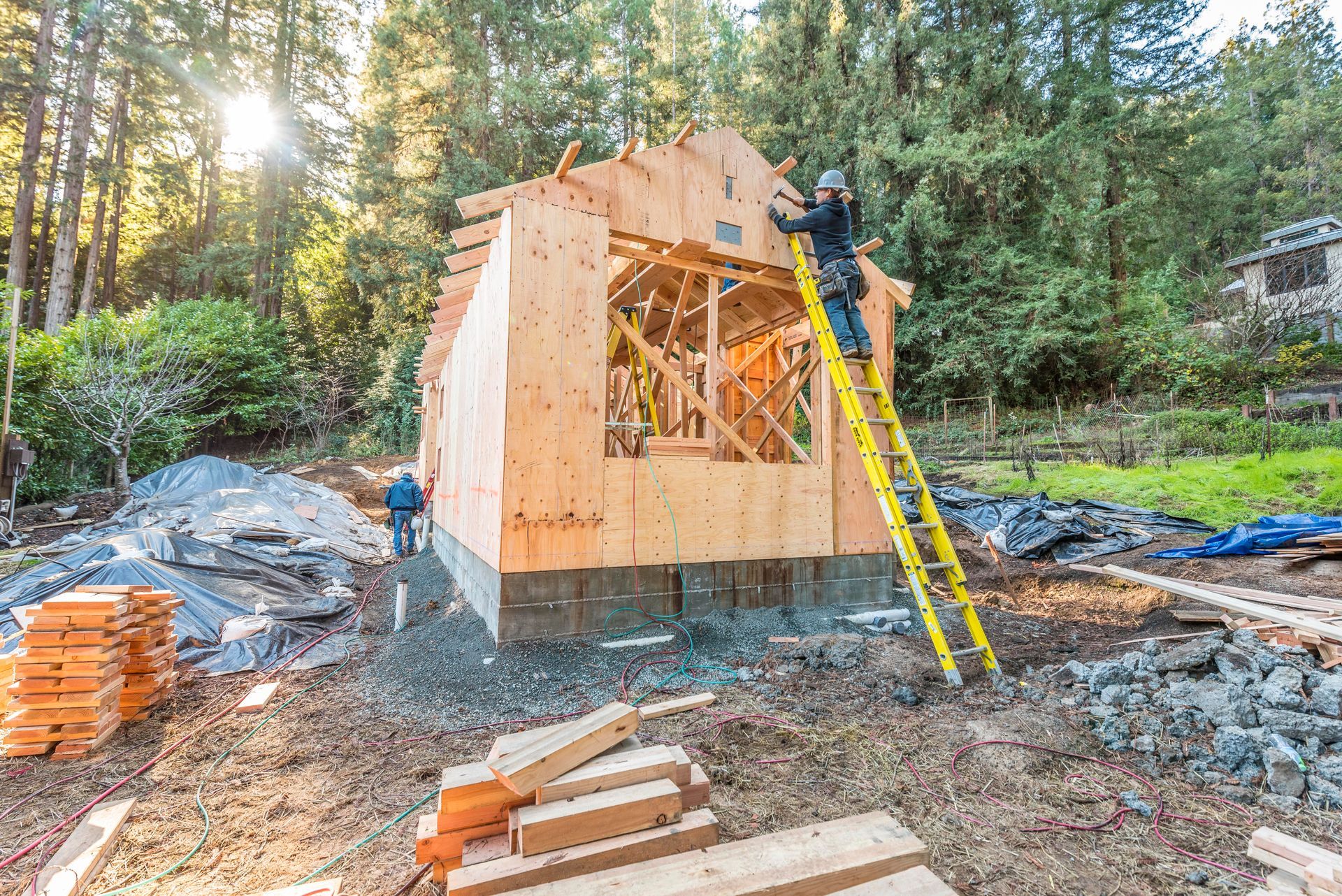 Wooden house under construction in a forest, with a worker on a yellow ladder and building materials nearby