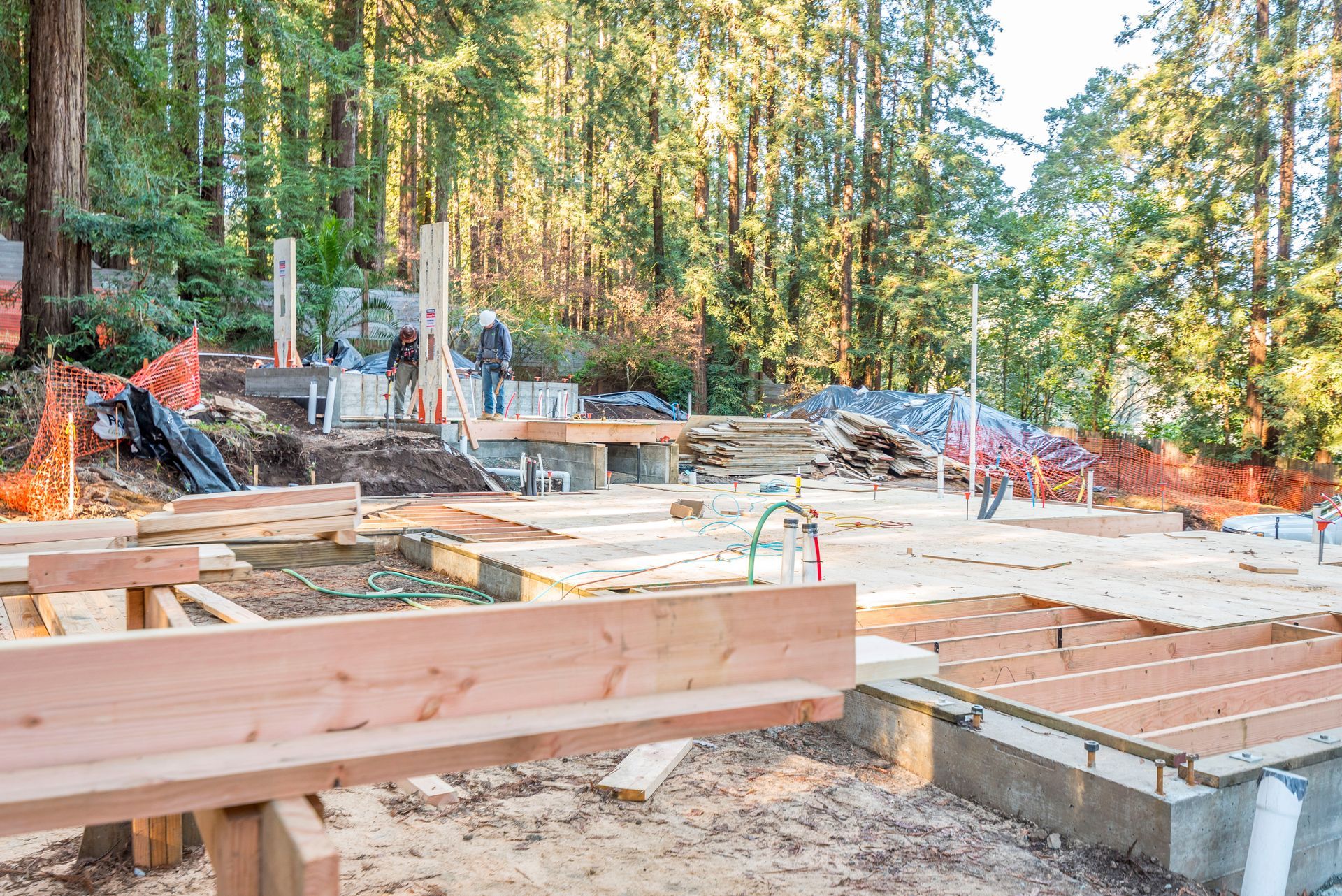 Construction site with concrete foundation, lumber framing, and orange safety fencing in a wooded area