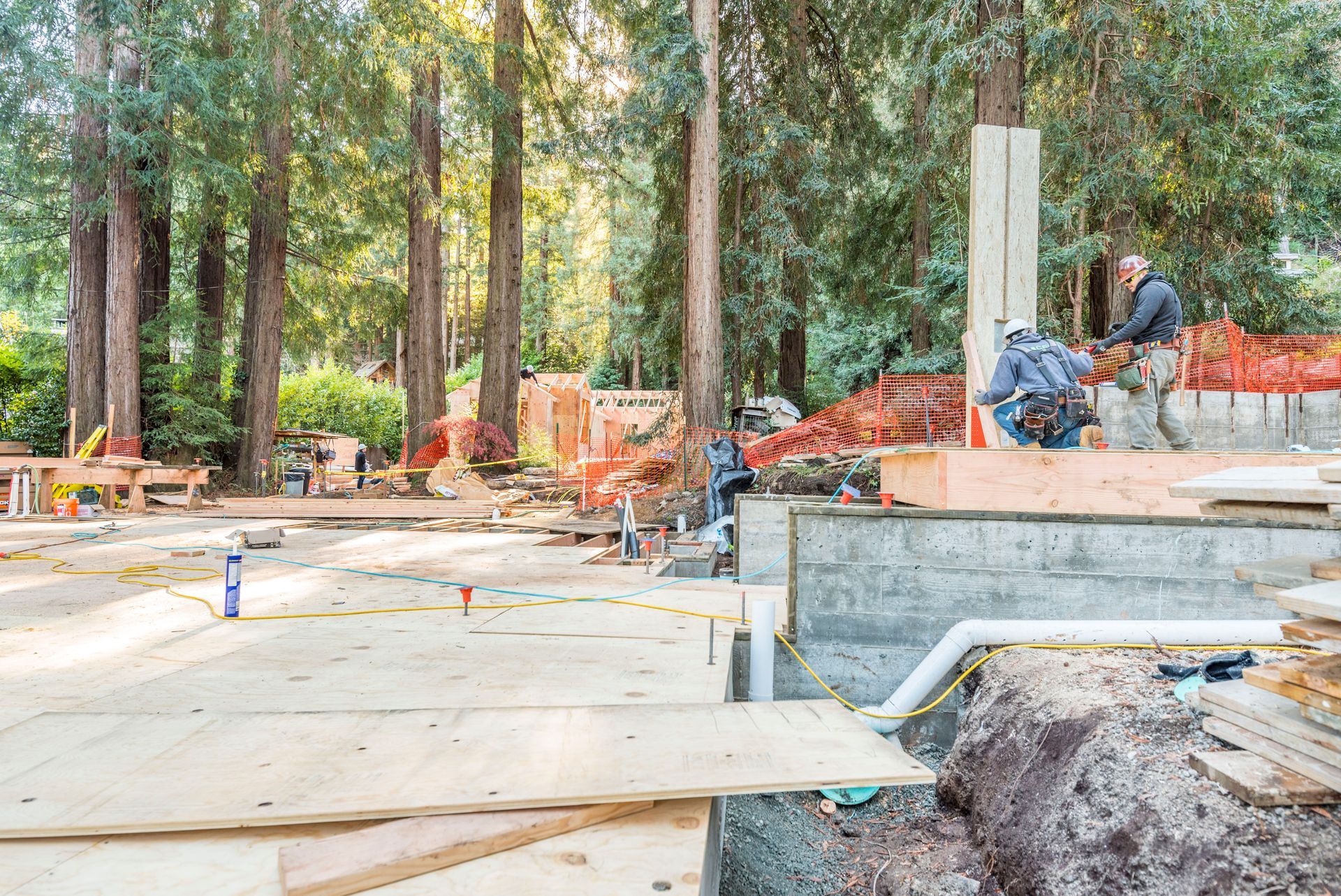 Construction site with concrete foundation, wooden forms, orange fencing, and workers among tall trees