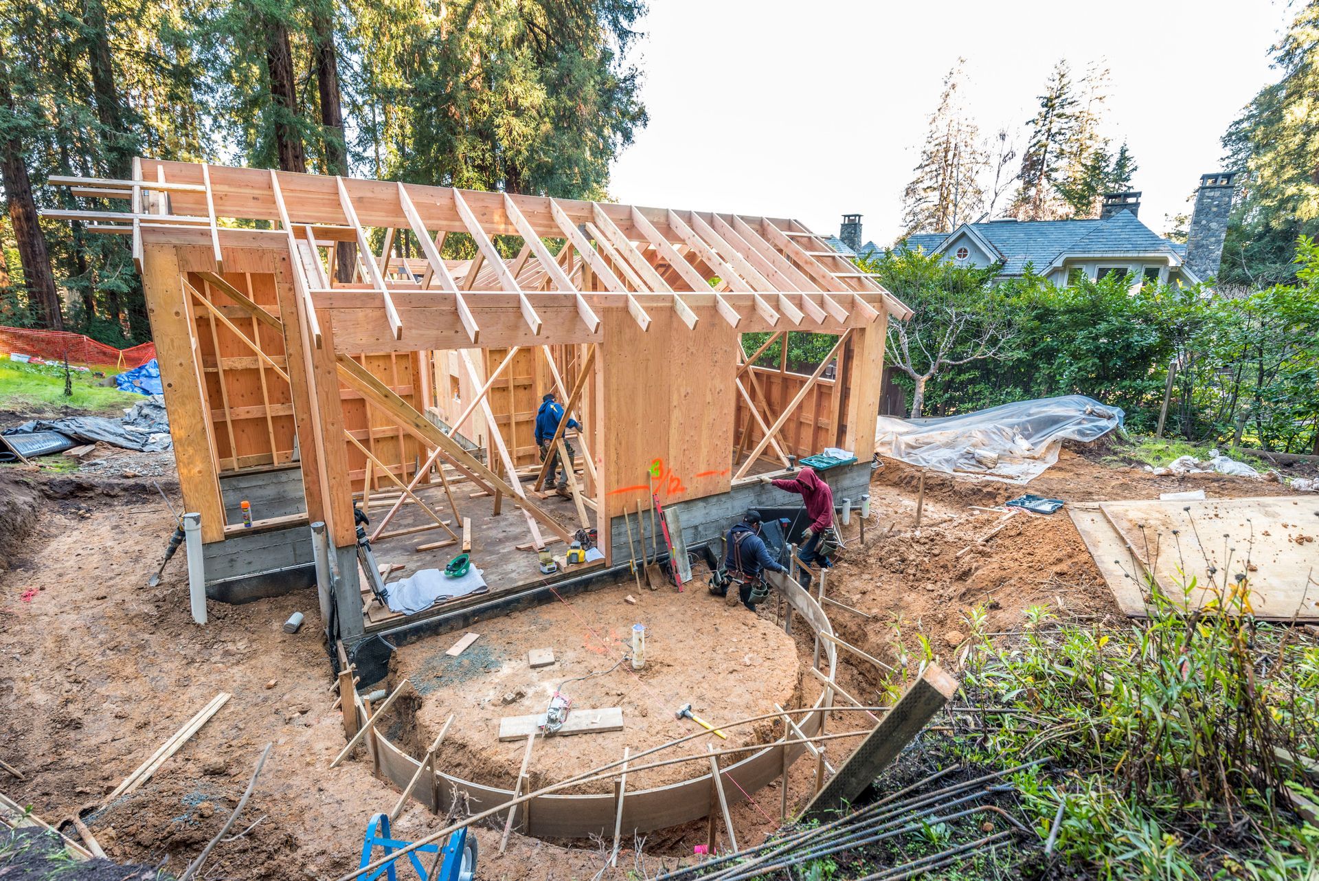Wood-framed small house under construction beside a pond, with workers and building materials around it