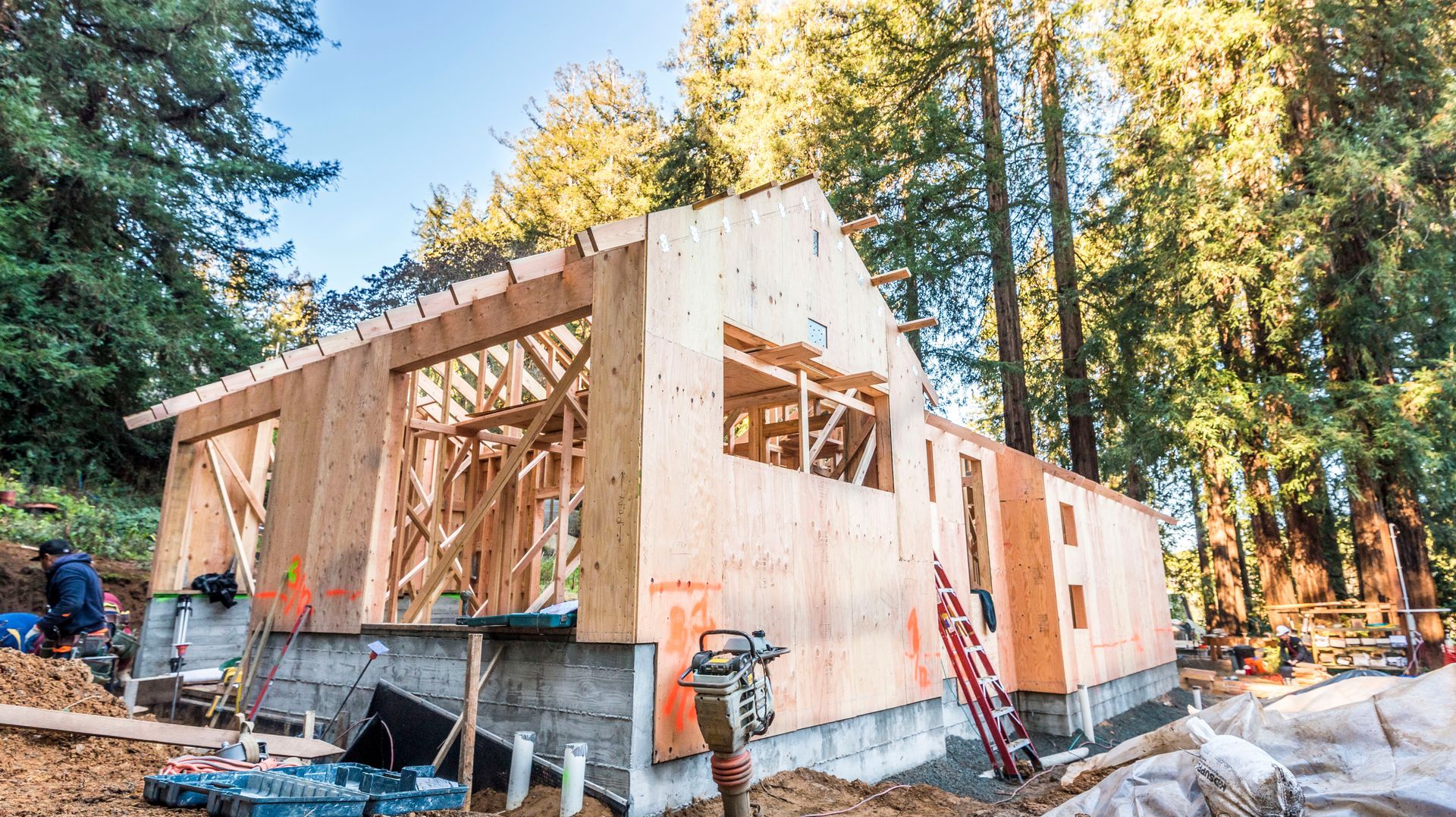 Wood-frame house under construction with exposed studs and tall trees in the background