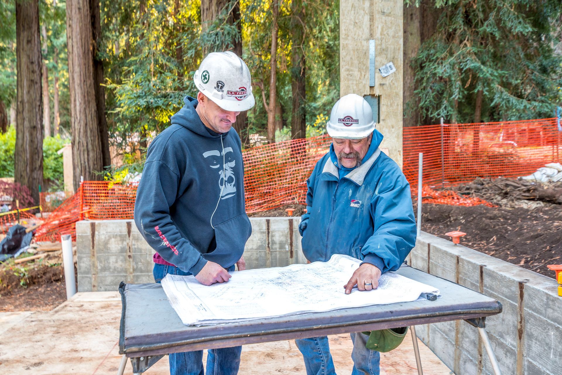 Two workers in hard hats review a map on a table at a construction site.
