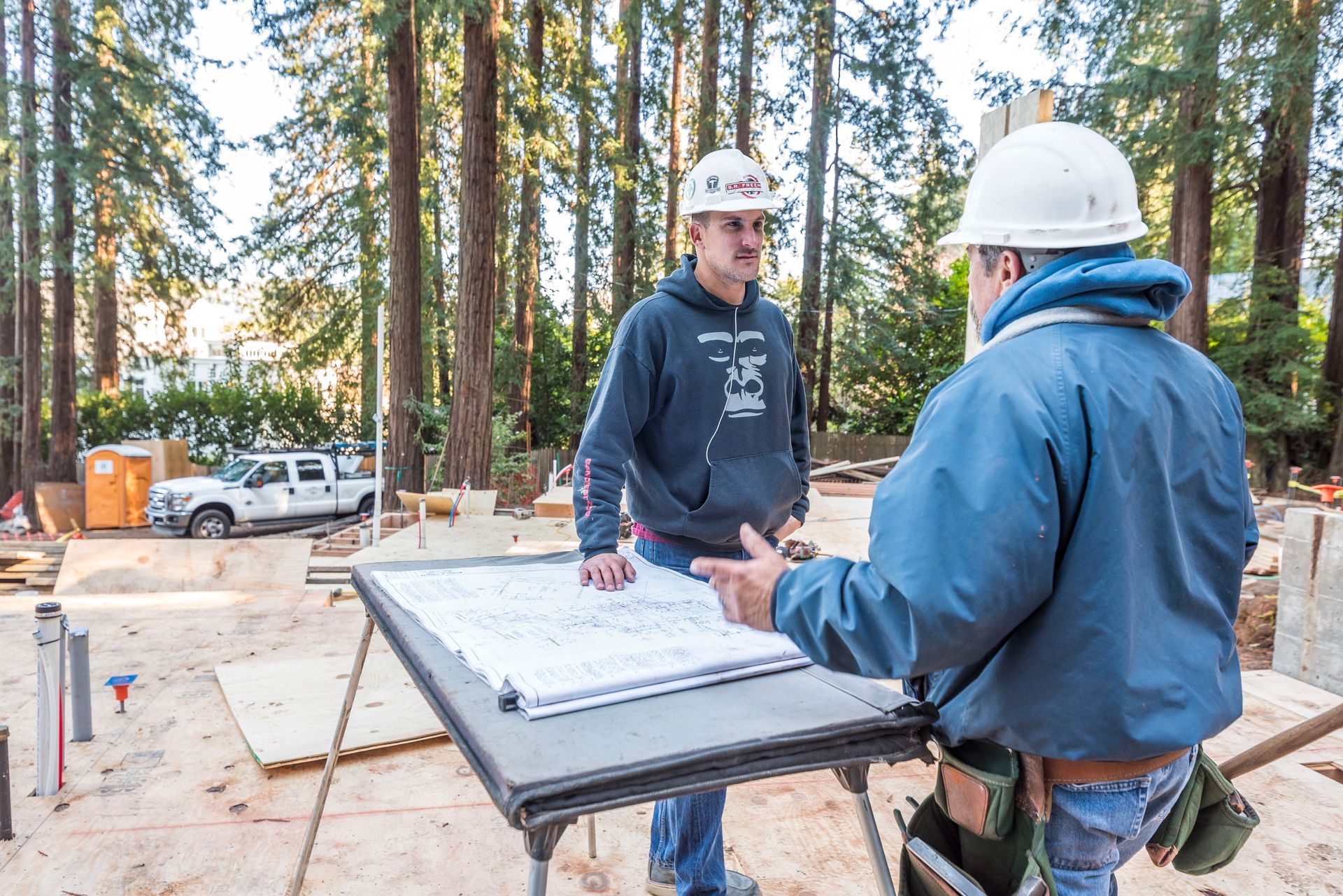 Two construction workers in hard hats reviewing plans at a wooded building site