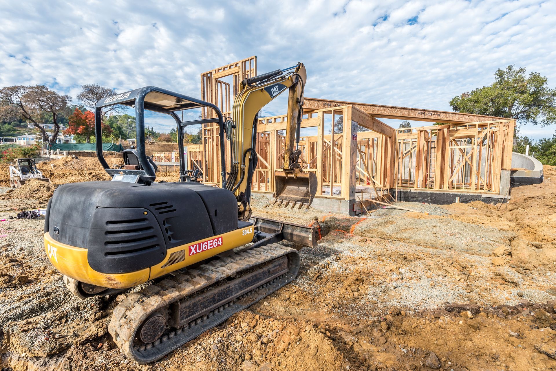 Excavator at a construction site beside a partially framed wooden building under a blue sky