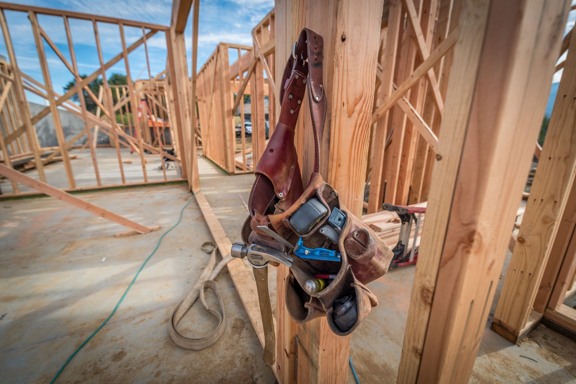 Worker hanging upside down from wood framing inside a house under construction, holding a tool belt.