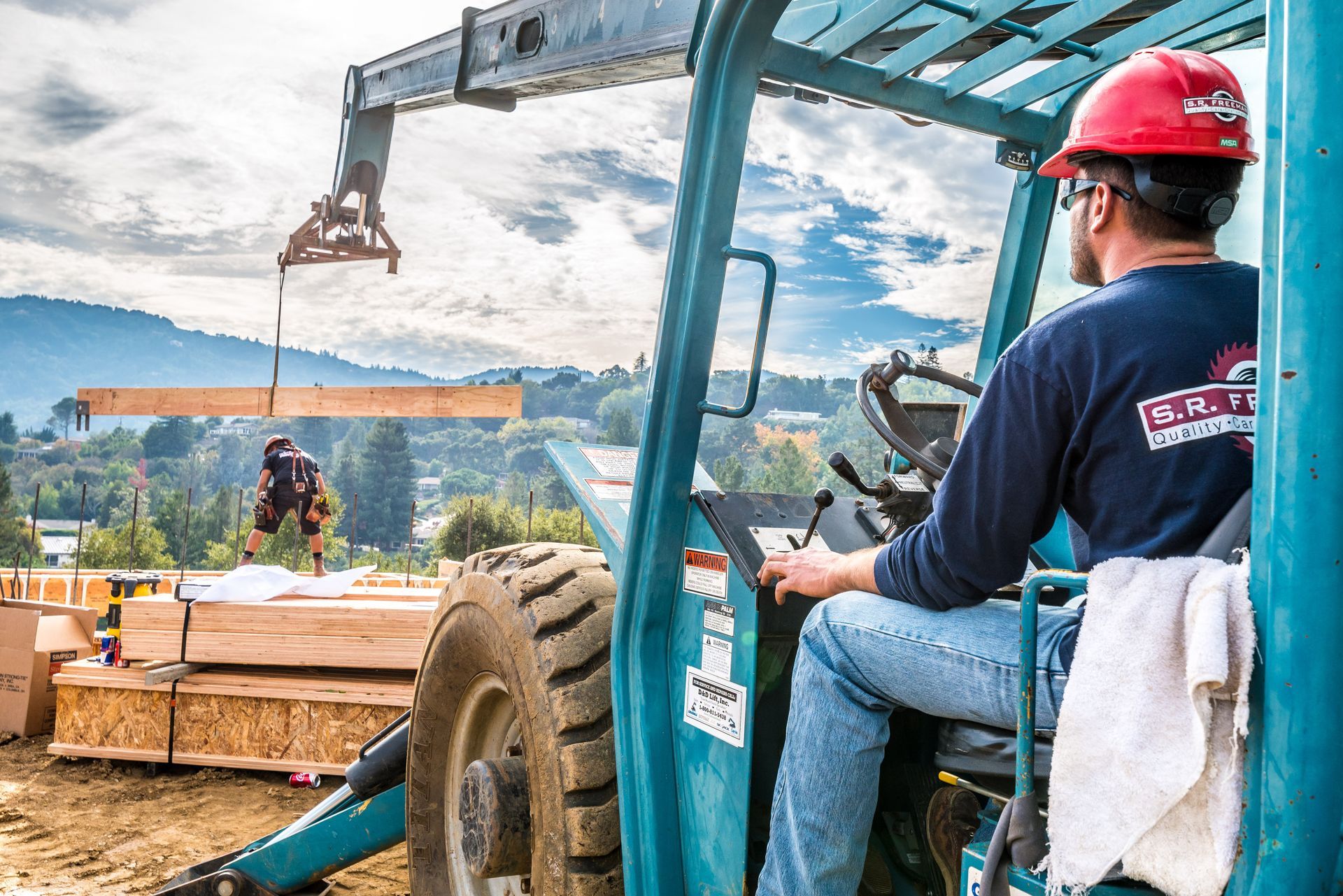 Worker in a red hard hat operating a forklift at a construction site, moving wooden materials.