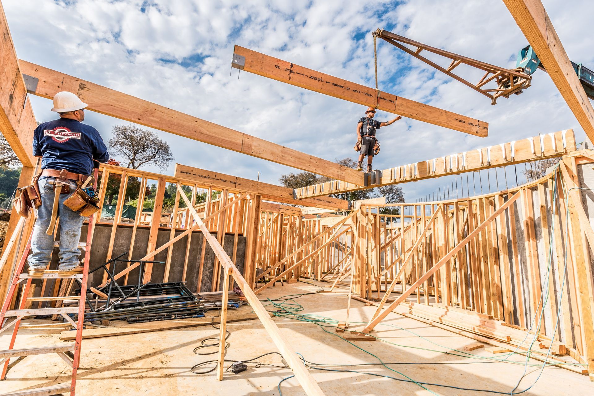 Construction workers framing a wooden house under a blue sky, with roof beams overhead.