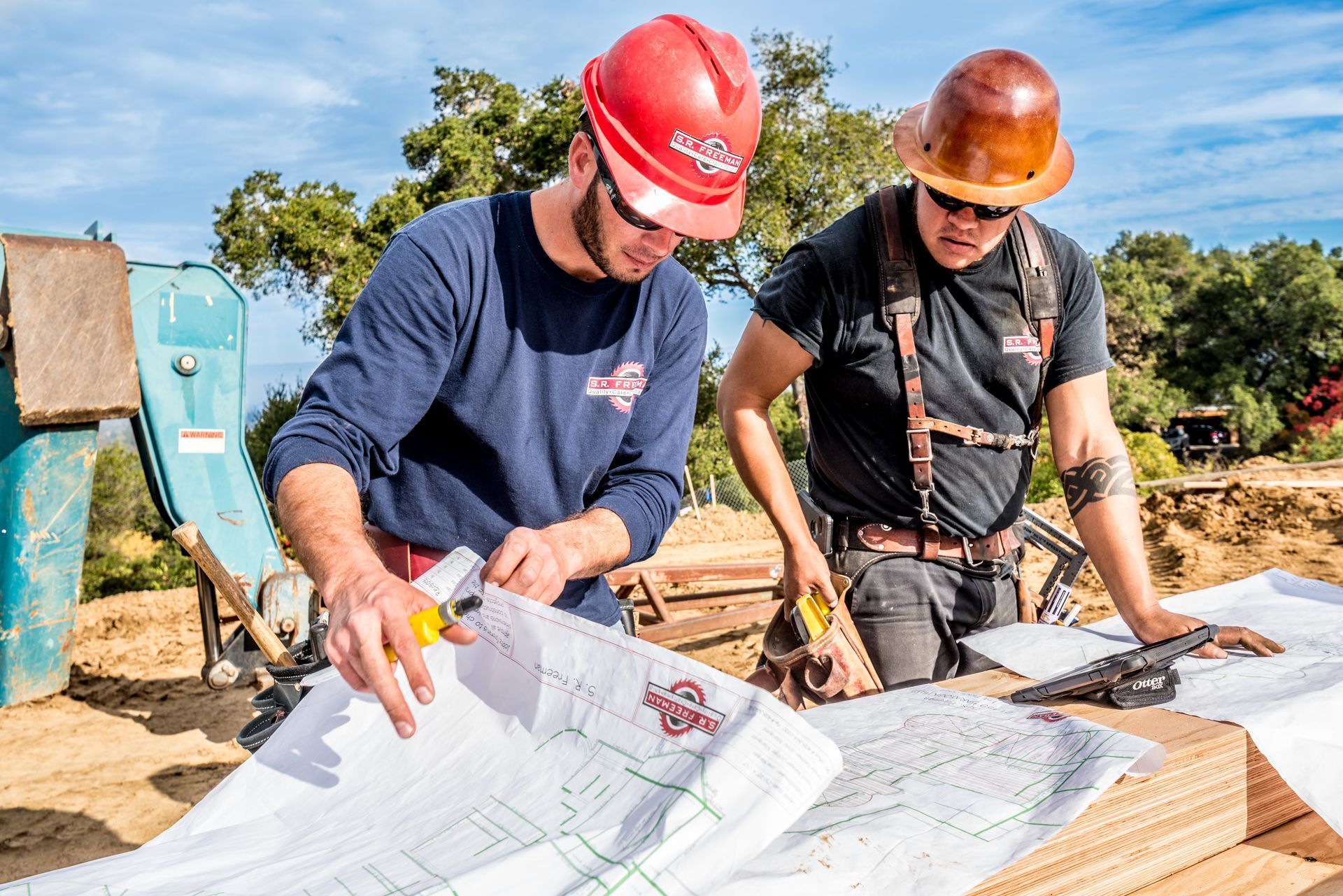 Two workers in hard hats review blueprints at a construction site outdoors.
