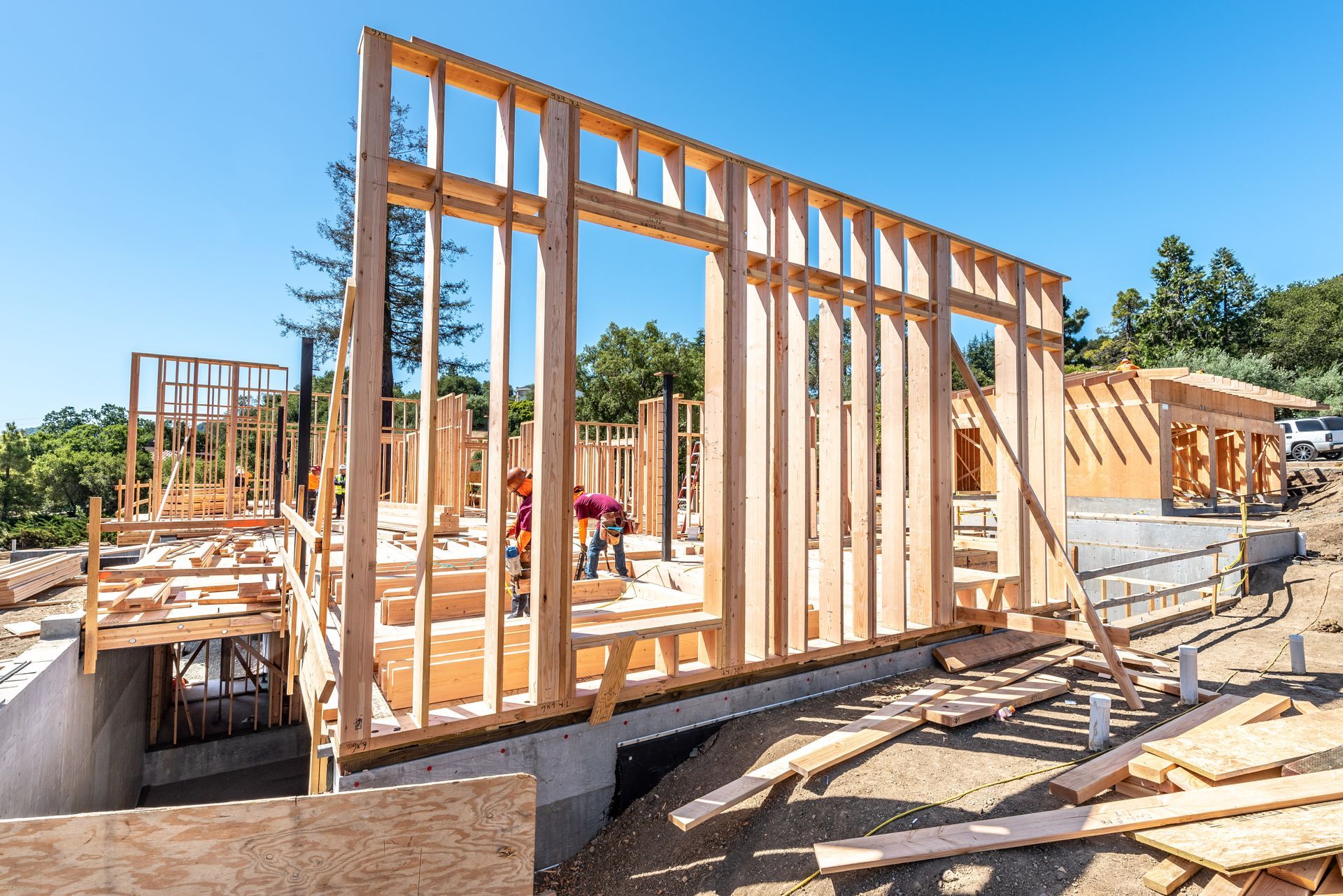 Wood-framed house under construction with workers and stacks of lumber on a sunny day